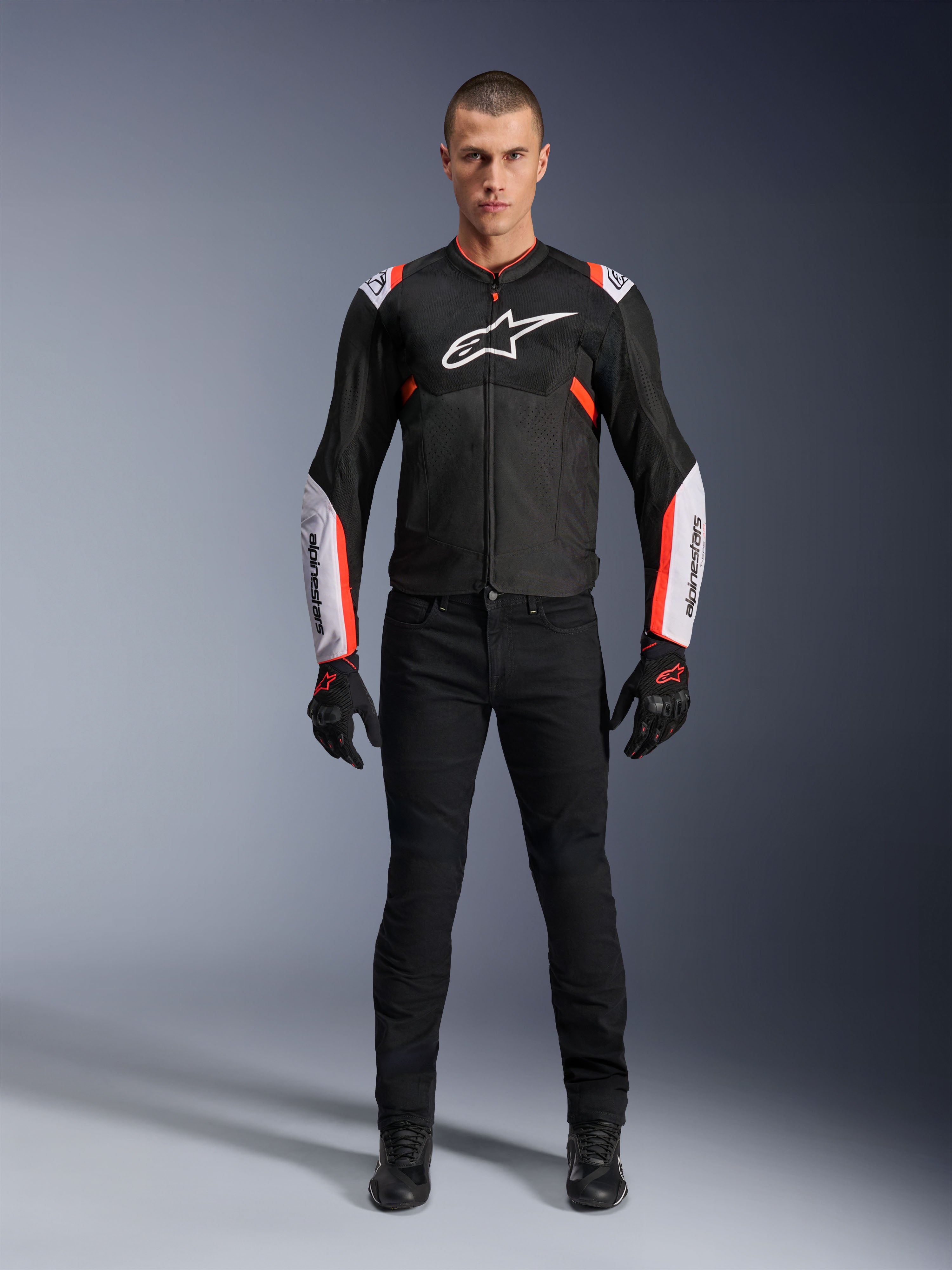 Modello che indossa la giacca Alpinestars T-Sps Air V2 in Black White Red Fluo, caratterizzata da ampi pannelli in mesh tecnico e logo bianco su petto e maniche, abbinata a pantaloni tecnici da guida e guanti neri, abbigliamento da moto sportivo urbano