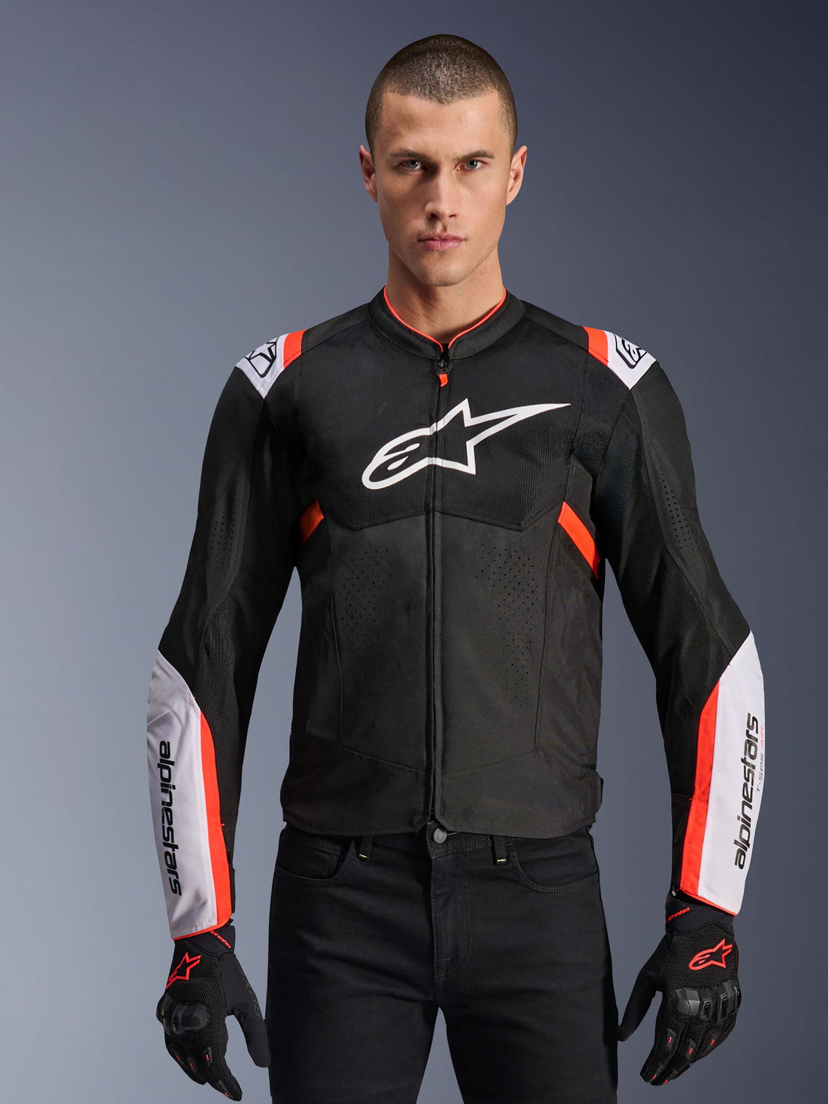 Alpinestars T-Sps Air V2 Jacket, giacca da moto sportiva, nero con accenti bianco e rosso fluo, uomo che indossa un design traforato in mesh con grande logo sul petto e maniche con marchio, abbinata a guanti e pantaloni neri