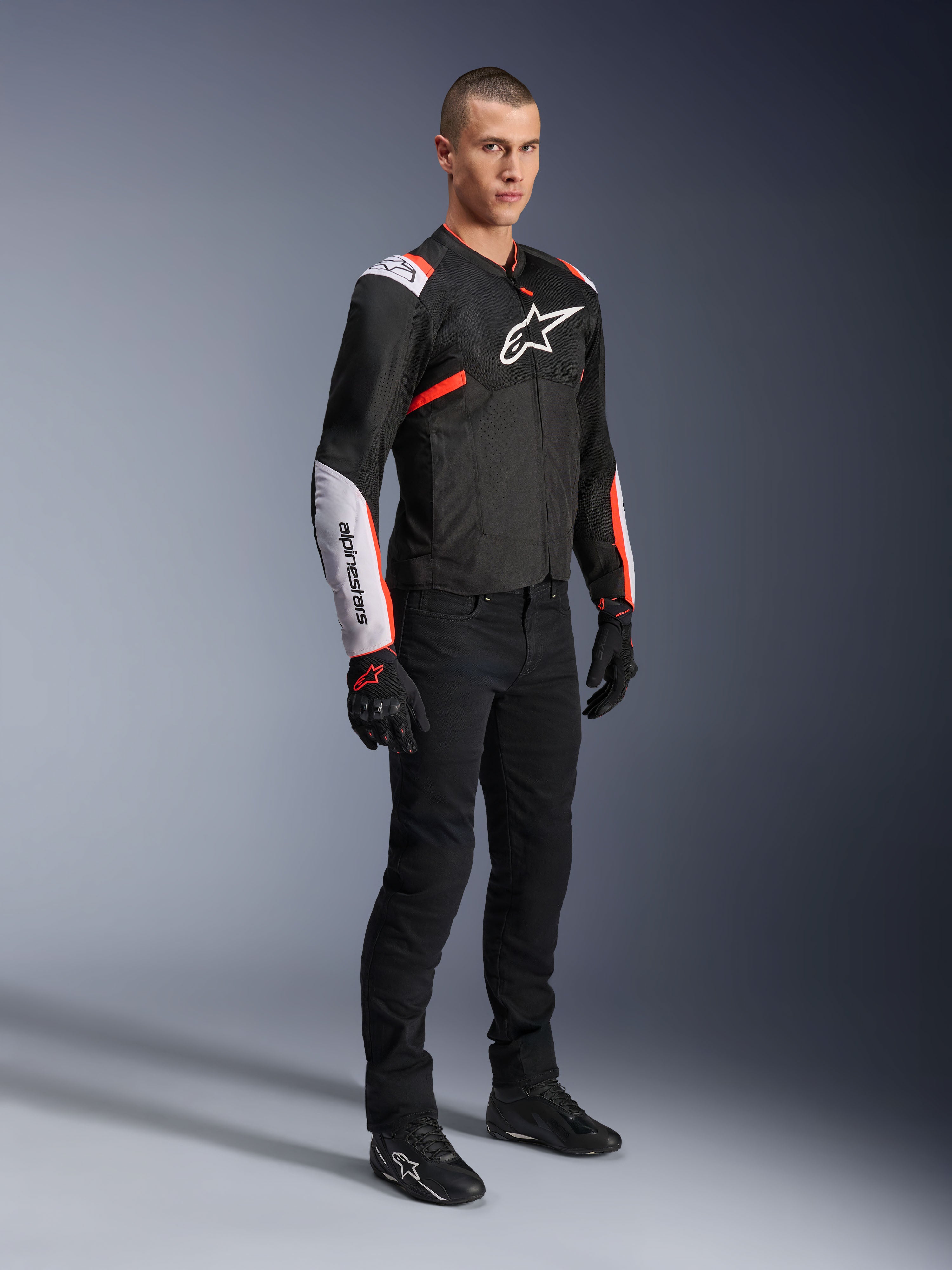 Giacca Alpinestars T-Sps Air V2, Nero Bianco Rosso Fluo, indossata da un modello uomo con pantaloni e guanti da moto neri, giacca da moto in tessuto con ampi pannelli in mesh, sezioni bianche su spalle e avambracci e logo Alpinestars sul petto.