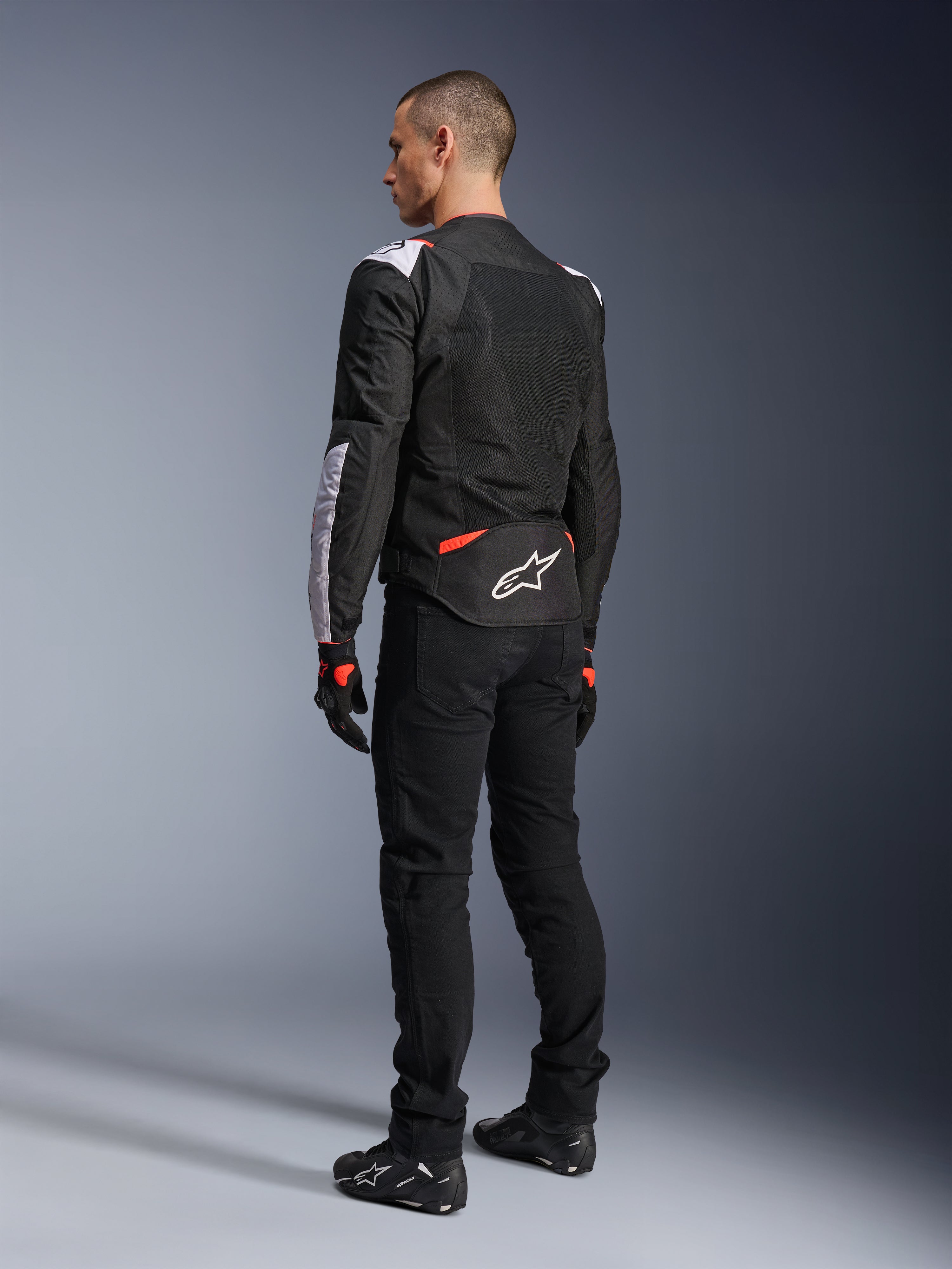 Giacca Alpinestars T-Sps Air V2, giacca da moto in tessuto, nero, bianco e rosso fluo, vista posteriore su modello, pannelli in mesh per la ventilazione, logo Astars bianco sulla parte inferiore della schiena, abbinata a pantaloni e guanti da moto neri