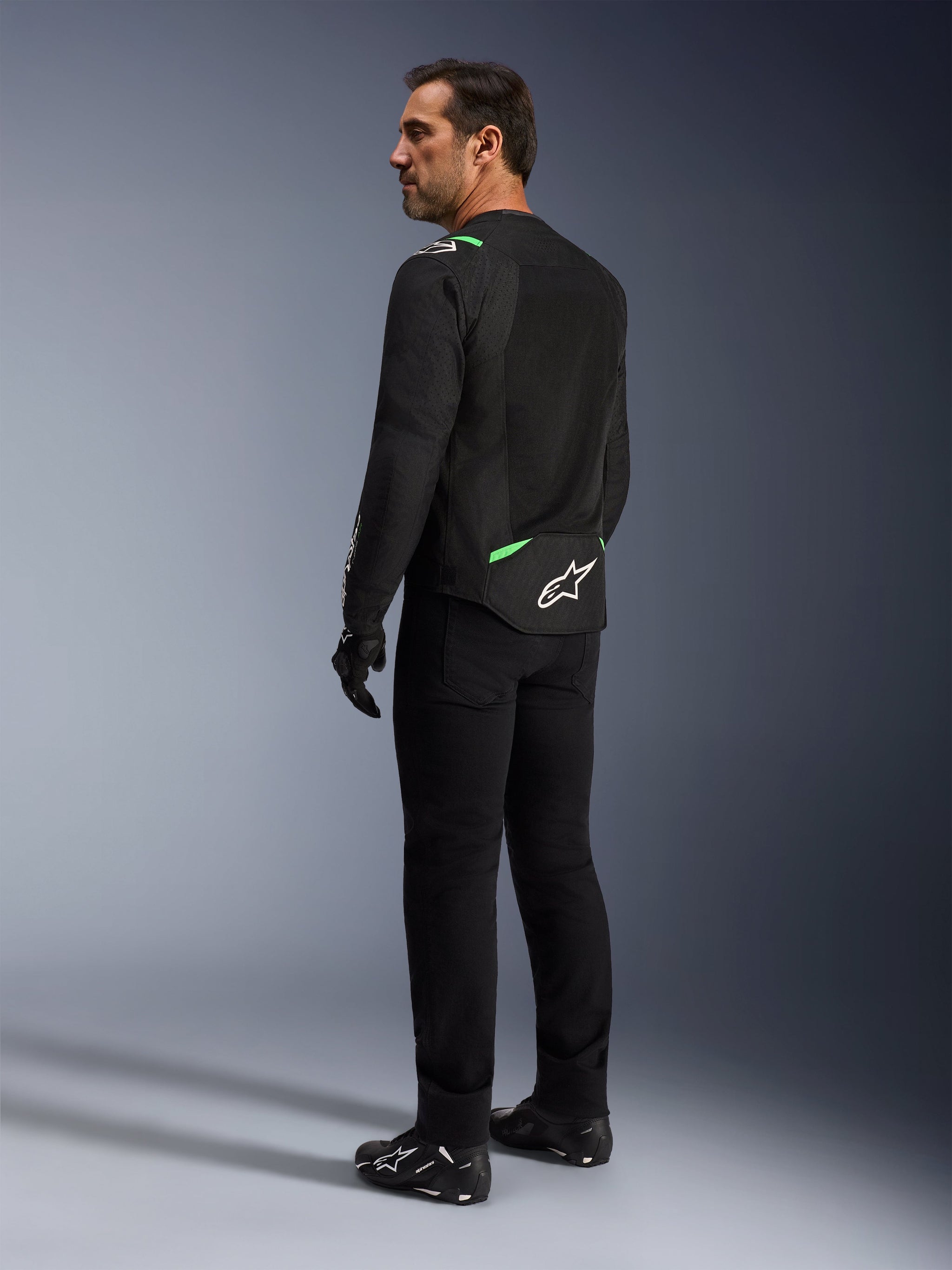 Giacca Alpinestars T-SPS Air V2 in Nero Verde Fluo indossata da un uomo, vista di profilo posteriore e laterale, abbinata a pantaloni da guida neri e scarpe da moto. La giacca presenta pannelli in rete tecnica ventilata, accenti verde fluo su spalle e parte bassa della schiena e il logo a stella bianco sul retro.