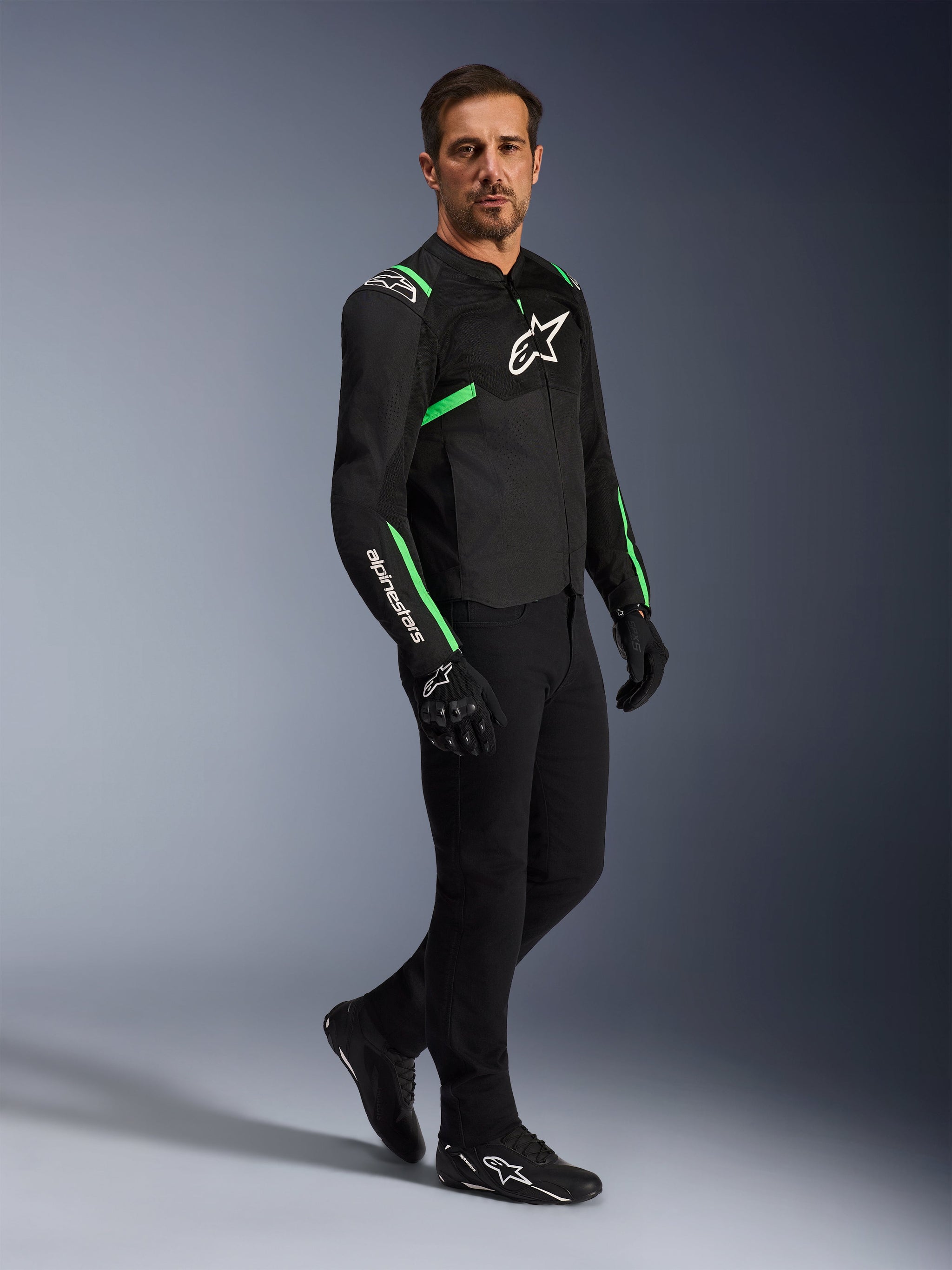 Modello che indossa la giacca Alpinestars T-SPS Air V2 in nero e verde fluo, abbinata a pantaloni e guanti da moto neri, giacca in tessuto dal taglio sportivo con pannelli in rete e accenti neon, outfit da moto urban completo