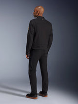 Giacca Alpinestars Bruiser, giacca da moto urban nera, vista posteriore su un modello, stile workwear classico, caratterizzata da pannelli plissettati sulle spalle e patch Oscar a spirale sulla manica, indossata con pantaloni neri e scarpe casual nere con suola color para.