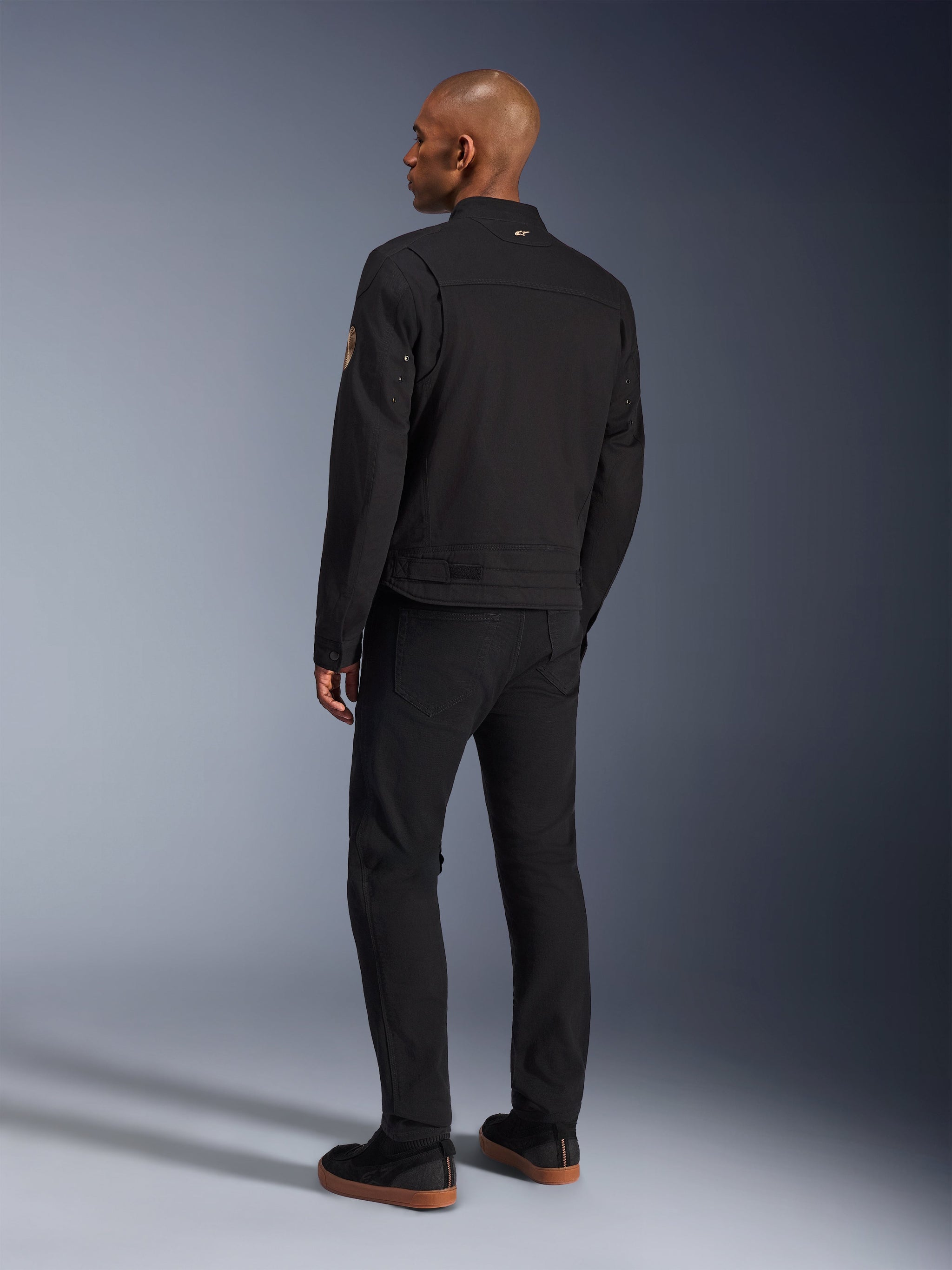 Giacca Alpinestars Bruiser, giacca da moto urban nera, vista posteriore su un modello, stile workwear classico, caratterizzata da pannelli plissettati sulle spalle e patch Oscar a spirale sulla manica, indossata con pantaloni neri e scarpe casual nere con suola color para.