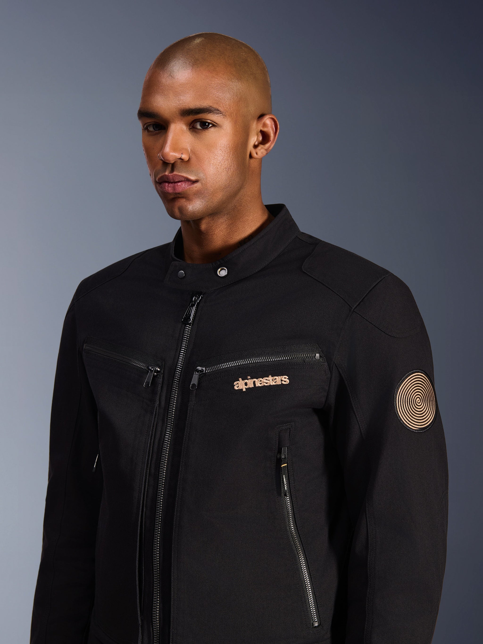 Modello che indossa la Giacca Alpinestars Bruiser, nera, giacca da moto urbana in twill di policotone, con tasche con cerniera, logo ricamato in oro sul petto e patch a spirale Oscar sulla manica, stile moderno per il commuting