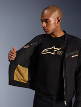 Giacca Alpinestars Bruiser, giacca da moto urbana nera, vista ravvicinata che mostra l'interno aperto con tasca in rete dorata e il branding 'Alpinestars Protects', indossata sopra una t-shirt nera con logo, abbigliamento da moto in tessuto con estetica workwear
