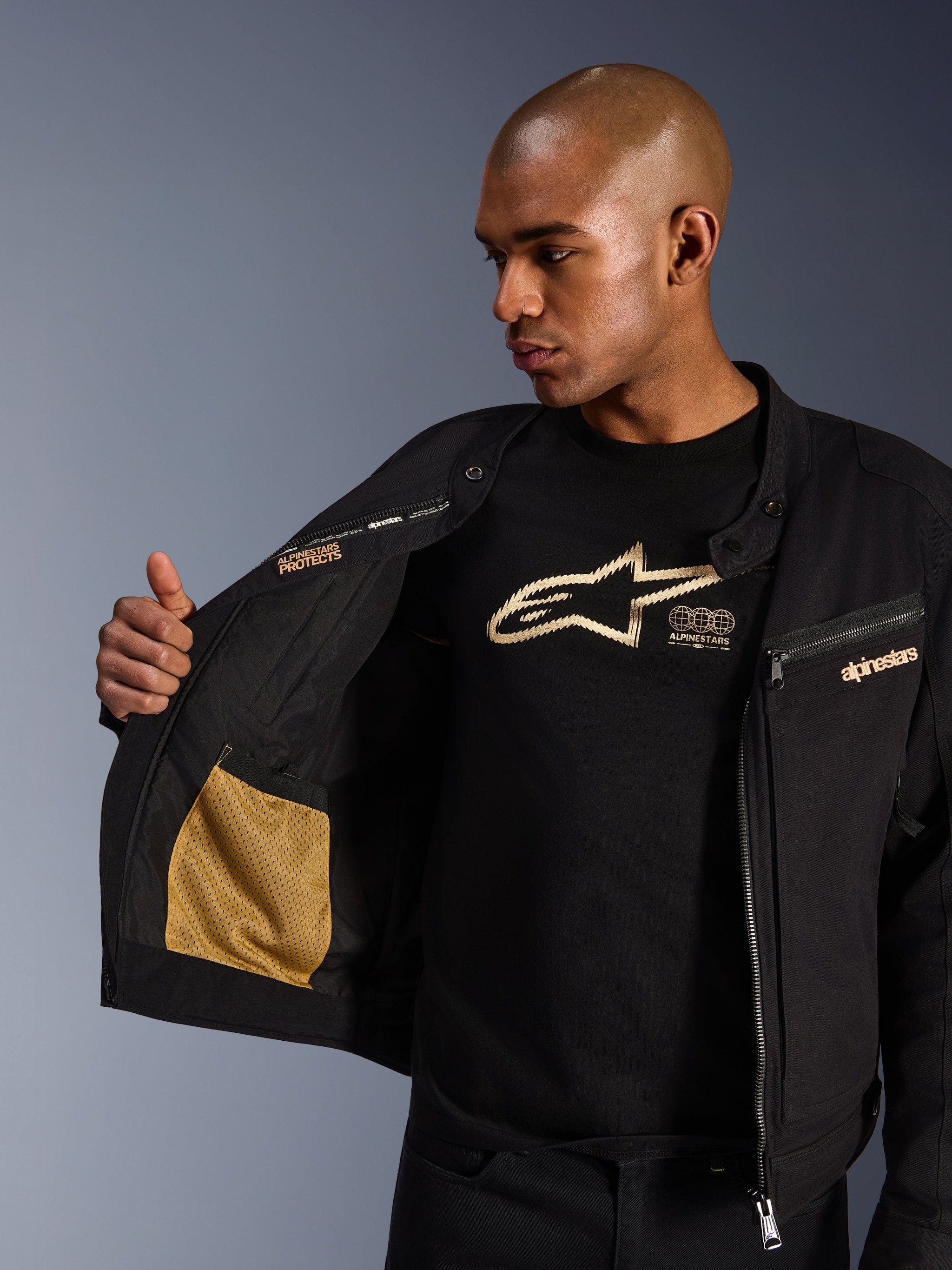 Giacca Alpinestars Bruiser, giacca da moto urbana nera, vista ravvicinata che mostra l'interno aperto con tasca in rete dorata e il branding 'Alpinestars Protects', indossata sopra una t-shirt nera con logo, abbigliamento da moto in tessuto con estetica workwear