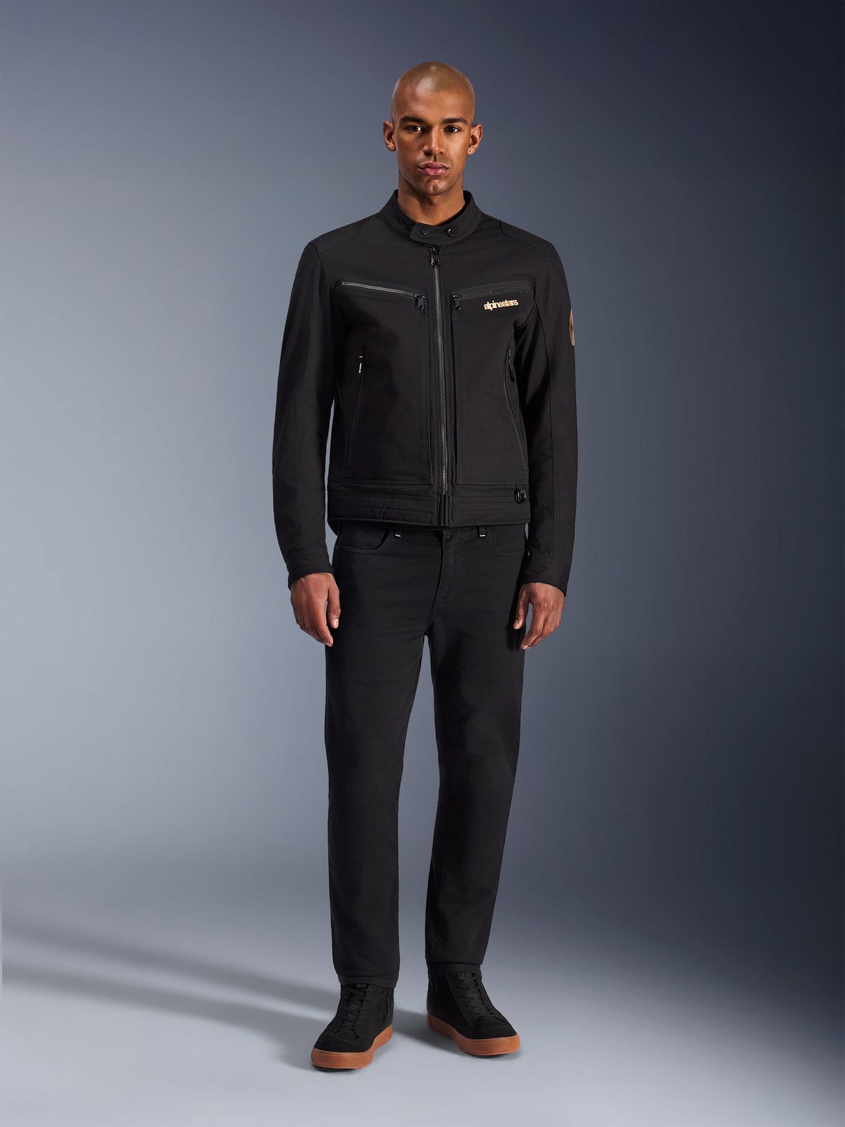 Giacca Alpinestars Bruiser, giacca da moto urbana nera, indossata da un modello con pantaloni neri e sneakers alte, vista frontale con tasche pettorali con cerniera e logo ricamato, design classico di ispirazione workwear per gli spostamenti quotidiani