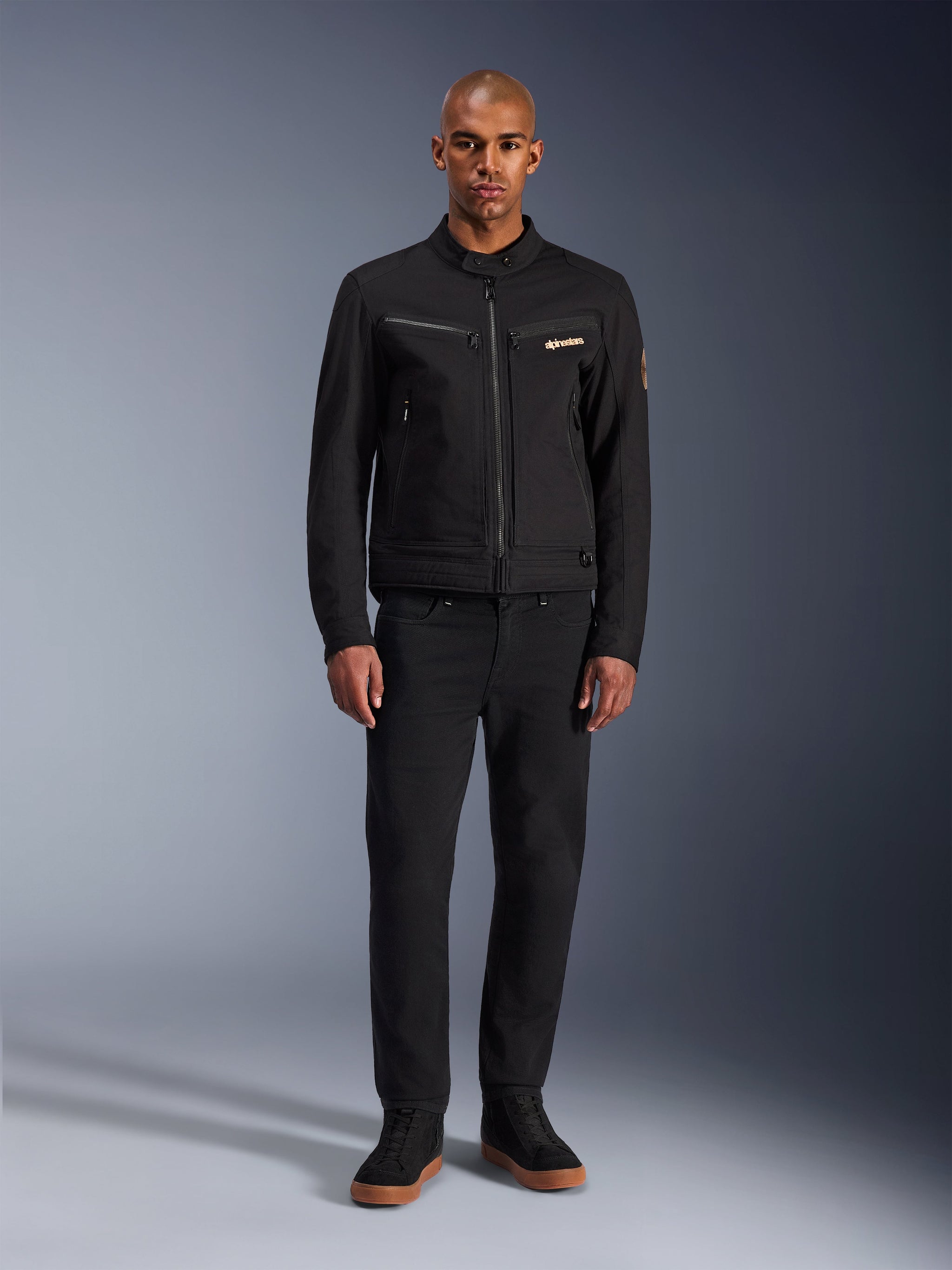 Giacca Alpinestars Bruiser, giacca da moto urbana nera, indossata da un modello con pantaloni neri e sneakers alte, vista frontale con tasche pettorali con cerniera e logo ricamato, design classico di ispirazione workwear per gli spostamenti quotidiani