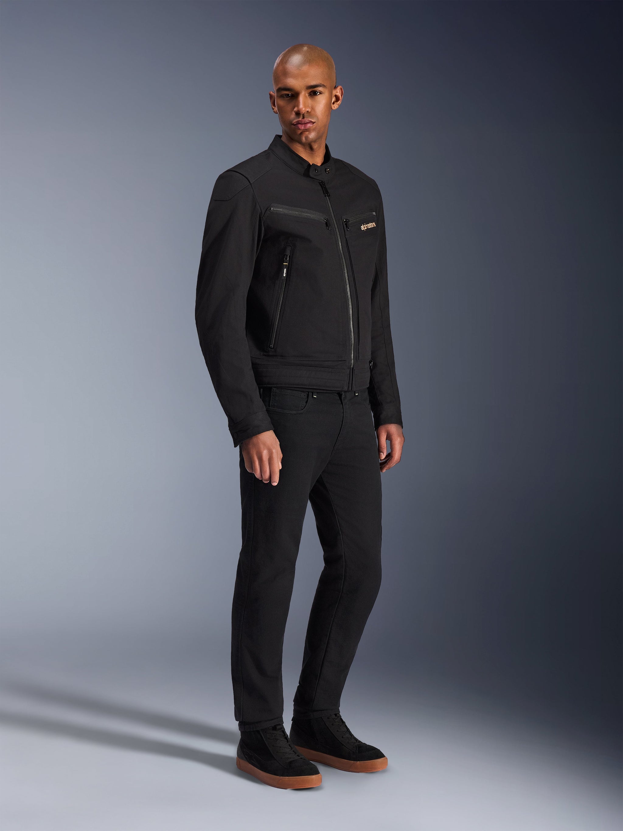 Giacca Alpinestars Bruiser, giacca da moto urbana, nero, indossata da un modello con pantaloni neri e sneakers, dotata di tasche sul petto con cerniera, tessuto twill di policotone e un design elegante ispirato al workwear per la guida urbana
