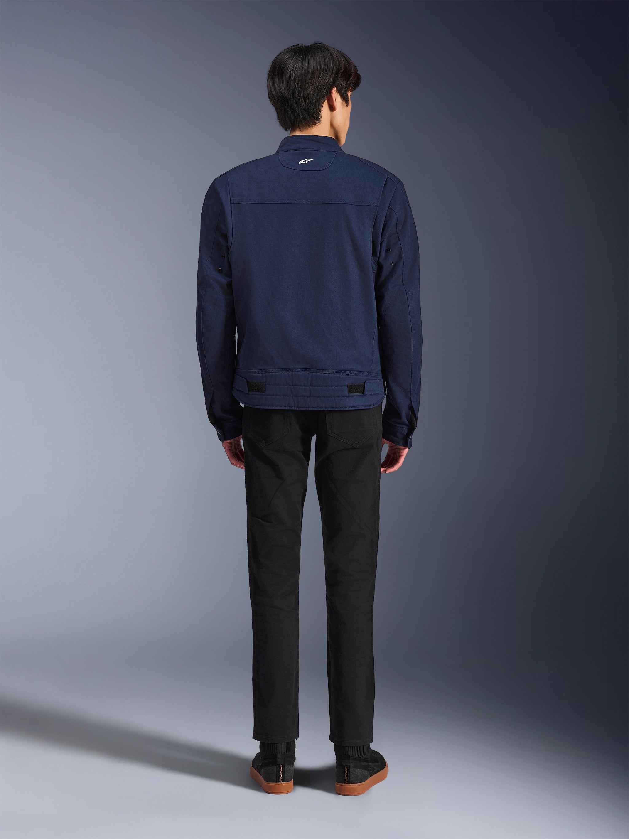 Giacca Alpinestars Bruiser in blu scuro, vista posteriore su modello, giacca da moto in tessuto dal design ispirato al workwear, con un piccolo logo bianco sul colletto posteriore e linguette di regolazione in vita, abbinata a pantaloni neri