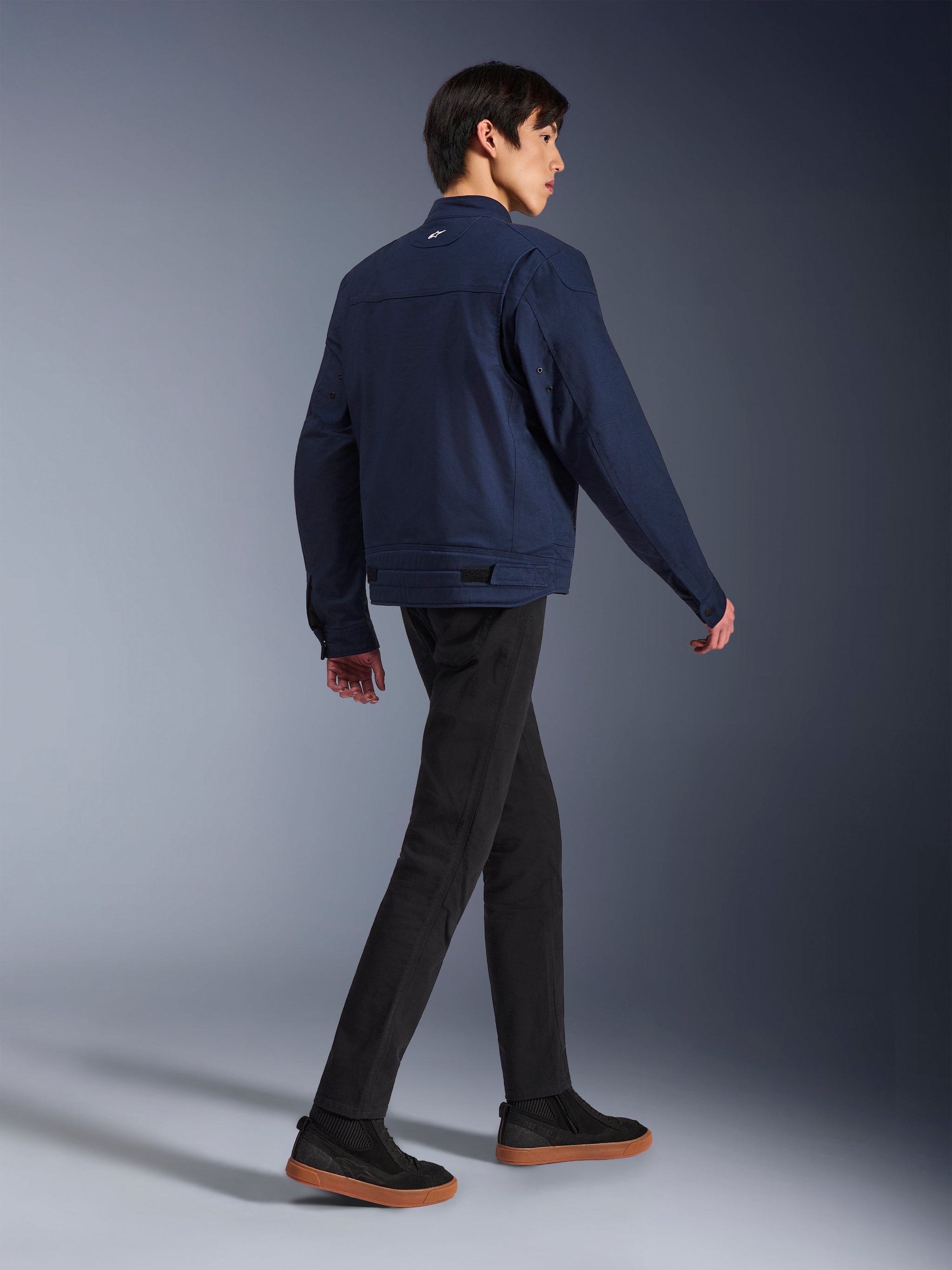 Giacca Alpinestars Bruiser in Dark Blue, giacca da moto urbana in tessuto, vista posteriore su modello che cammina, abbinata a pantaloni neri e sneakers con suola in gomma, con design ispirato al workwear e regolazioni in vita.