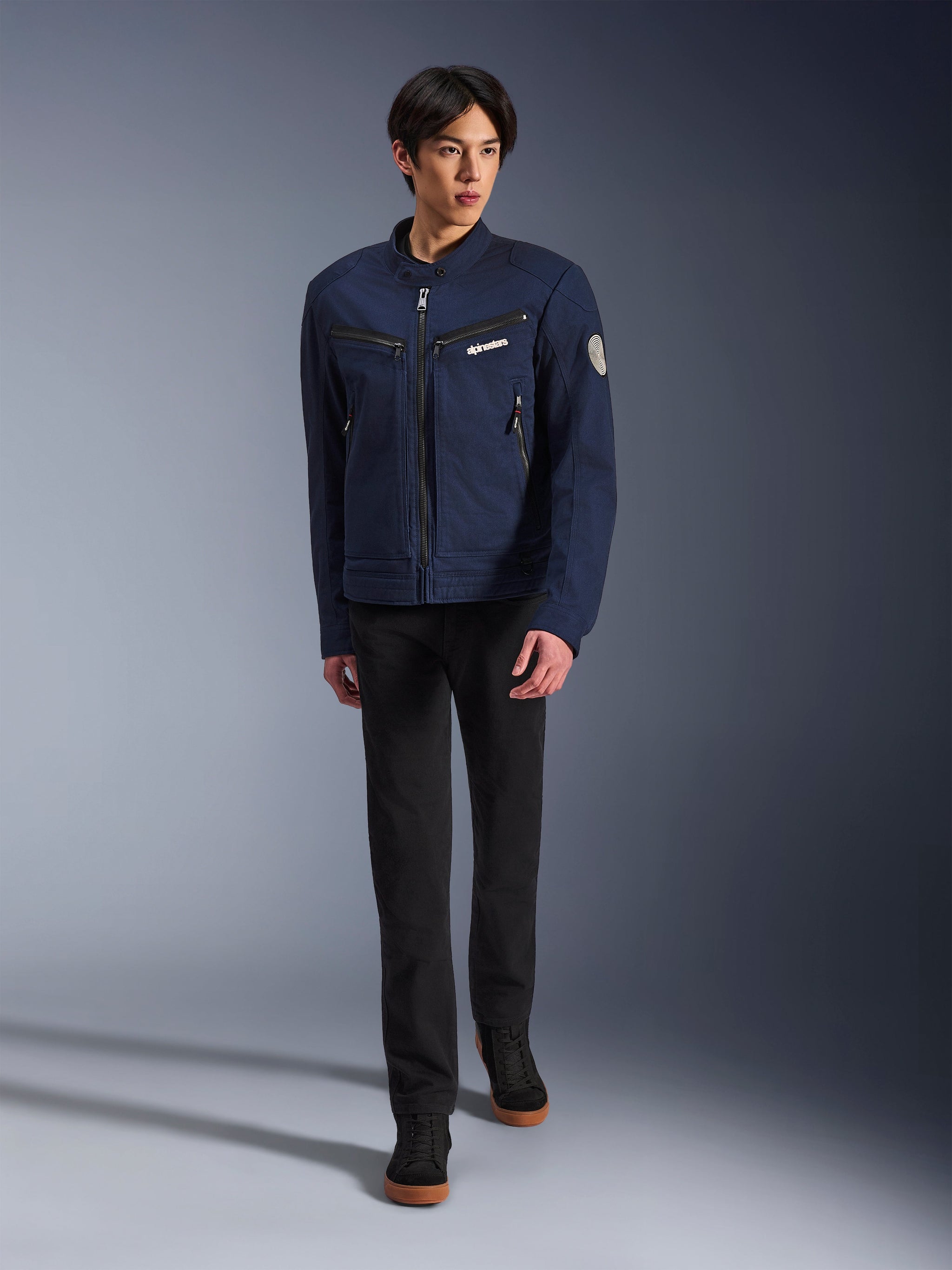 Modello con giacca Alpinestars Bruiser in Dark Blue, giacca da moto urbana in tessuto, abbinata a pantaloni neri e sneakers nere, estetica ispirata al workwear con tasche con zip e patch circolare sulla manica, streetwear contemporaneo per la guida in moto