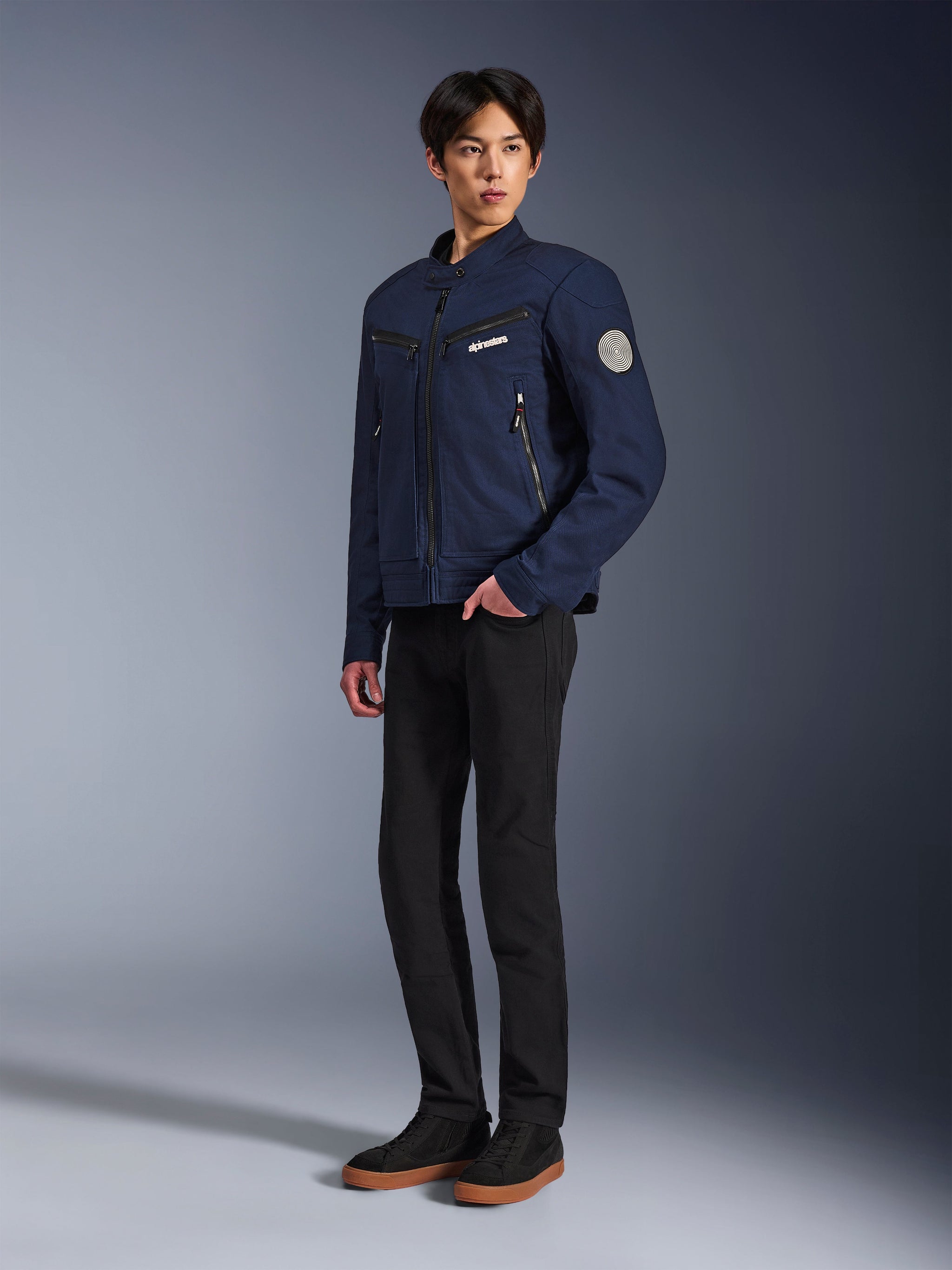 Giacca Alpinestars Bruiser in blu scuro, giacca da moto urbana in tessuto dal design d'ispirazione workwear, con tasche sul petto con cerniera e patch Oscar a spirale sulla manica, indossata con pantaloni neri e sneakers casual.