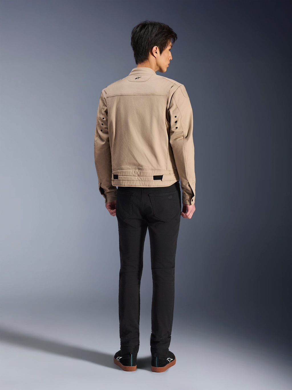Vista posteriore di un modello che indossa la giacca Alpinestars Bruiser in marrone, una giacca da moto urbana dal design ispirato all'estetica workwear, con dettagli di occhielli in metallo sulla schiena e regolatori in vita, abbinata a pantaloni neri e sneaker, stile street riding contemporaneo.