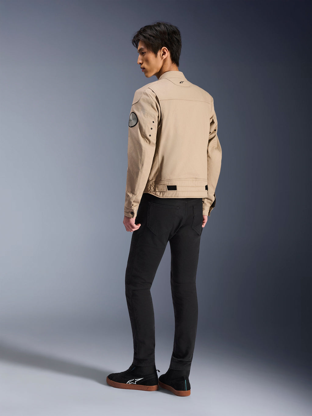 Modello maschile di spalle che indossa la giacca Alpinestars Bruiser color sabbia, giacca da moto urban contemporary in stile workwear, con patch circolare sulla manica, cinturini di regolazione in vita e spalle articolate, abbinata a pantaloni neri e sneakers nere.