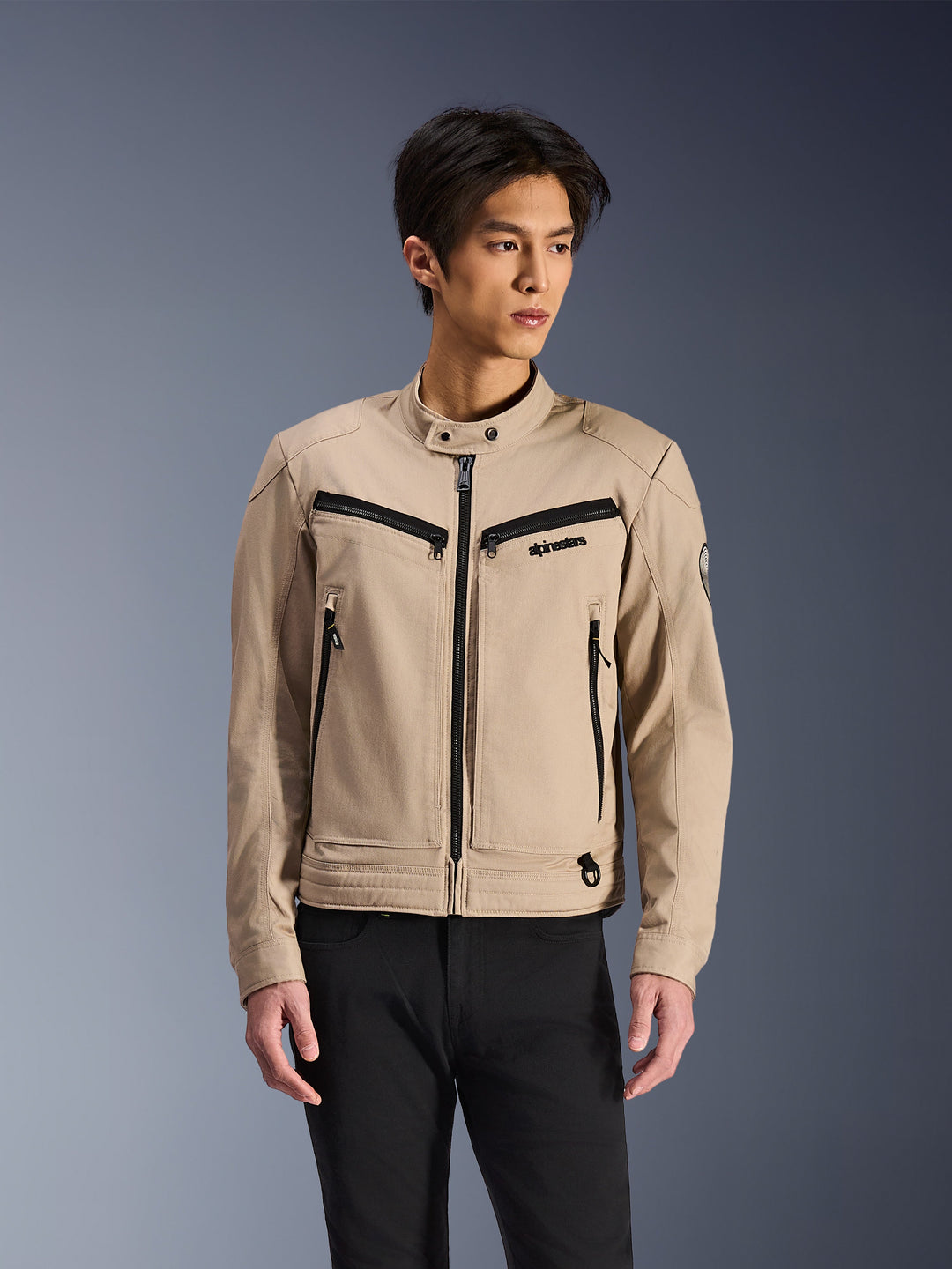 Alpinestars Bruiser Jacket, giacca da moto beige indossata da un modello uomo, dettagli con cerniere nere su petto e tasche laterali, colletto alto con bottone a pressione, abbigliamento da guida urbana ispirato al workwear, abbinata a pantaloni neri