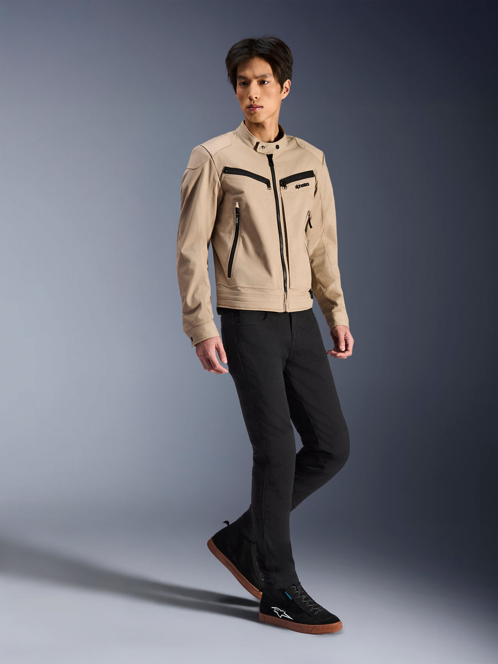 Giacca Alpinestars Bruiser, giacca da moto urbana, color tan, indossata da un modello uomo con pantaloni neri slim-fit e sneaker nere, costruzione in tessuto d'ispirazione workwear con accenti di cerniere nere, stile urban street contemporaneo.