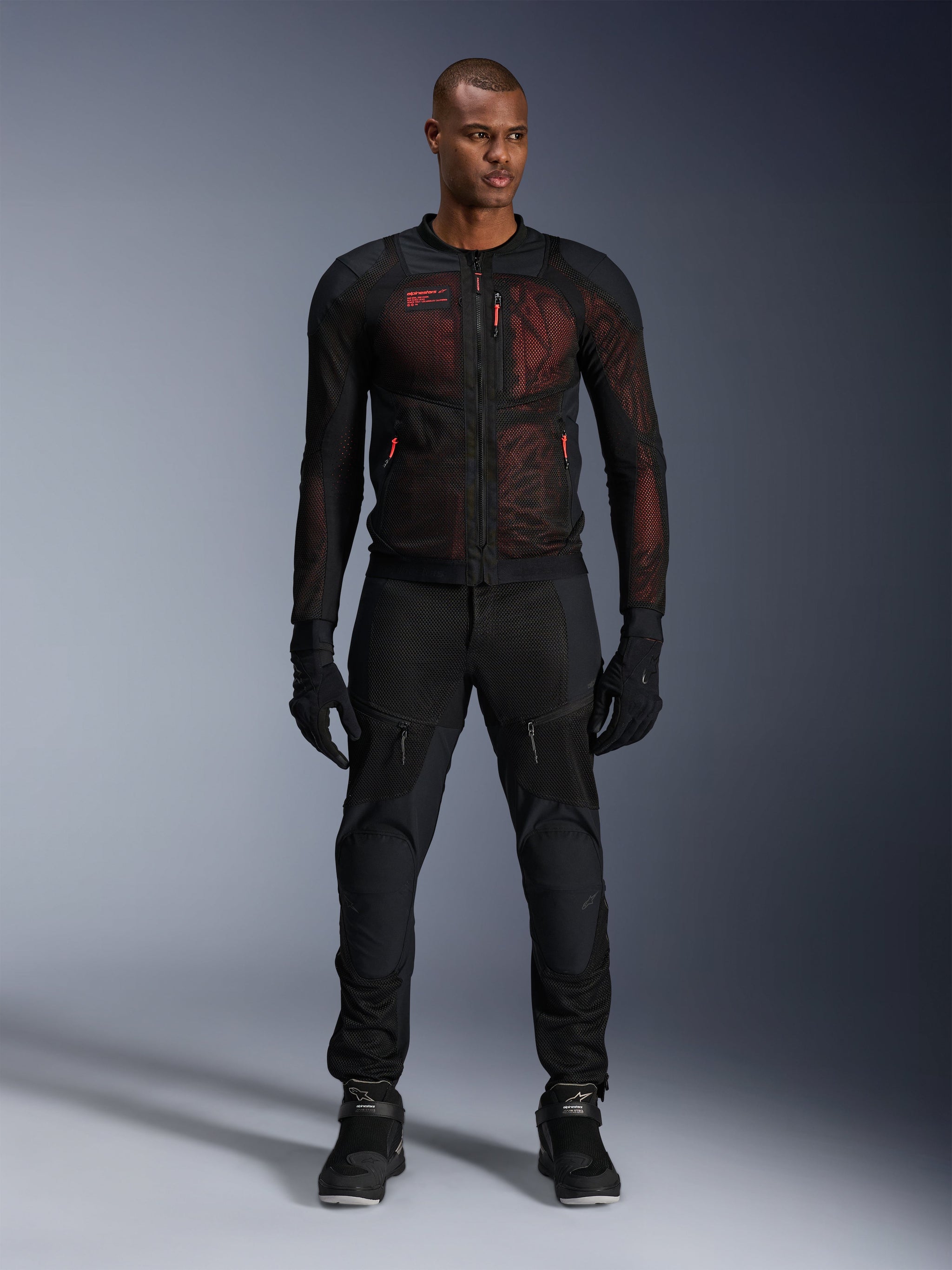 Giacca Alpinestars Troop-Air Pro, giacca tecnica traforata e corazzata, nero e rosso fluo, indossata da un modello con pantaloni e guanti coordinati, con pannelli in mesh ad alta ventilazione e accenti rossi, abbigliamento moto urban