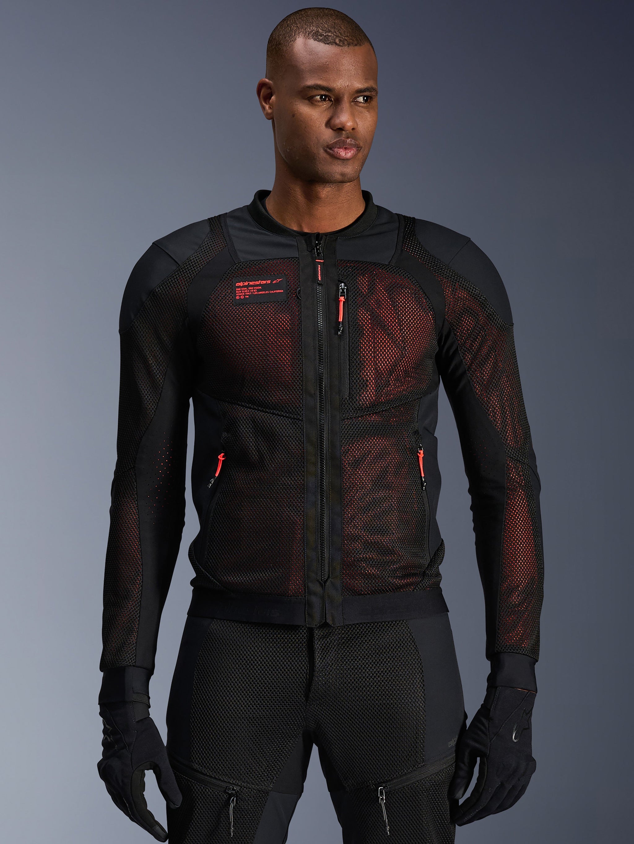 Giacca Alpinestars Troop-Air Pro, giacca da moto tecnica in rete corazzata in nero e rosso fluo, indossata da un modello, caratterizzata da ampi pannelli di ventilazione in rete, base rossa e tiranti delle cerniere rossi, stile urban riding.