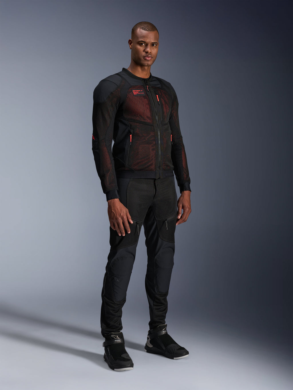 Giacca Alpinestars Troop-Air Pro in Nero Rosso Fluo, giacca da moto tecnica in mesh corazzata indossata da un modello uomo con pantaloni neri corazzati coordinati e stivali alti, ritratto in studio a figura intera.