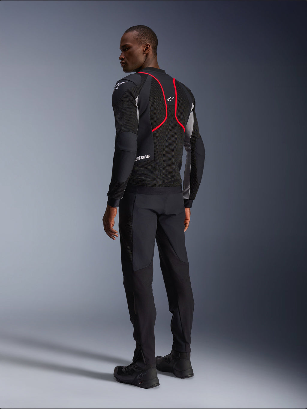 Alpinestars Troop-Air Pro Jacket, giacca da moto, nero, grigio cenere e rosso vivace, vista posteriore su modello, con linee d'accento rosse e pannelli di ventilazione in mesh, indossata con pantaloni e stivali neri, stile motociclistico urbano