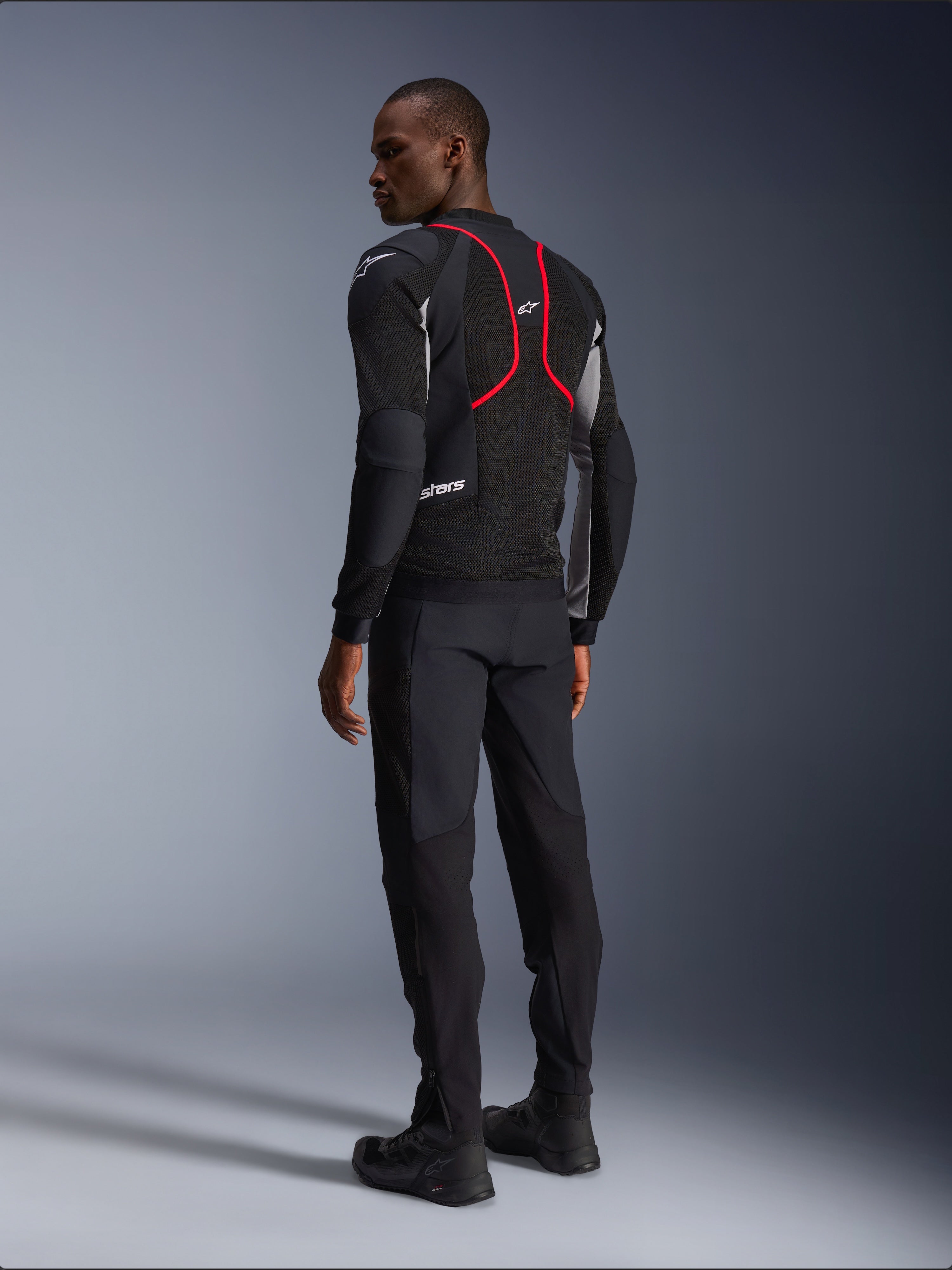 Alpinestars Troop-Air Pro Jacket, giacca da moto, nero, grigio cenere e rosso vivace, vista posteriore su modello, con linee d'accento rosse e pannelli di ventilazione in mesh, indossata con pantaloni e stivali neri, stile motociclistico urbano