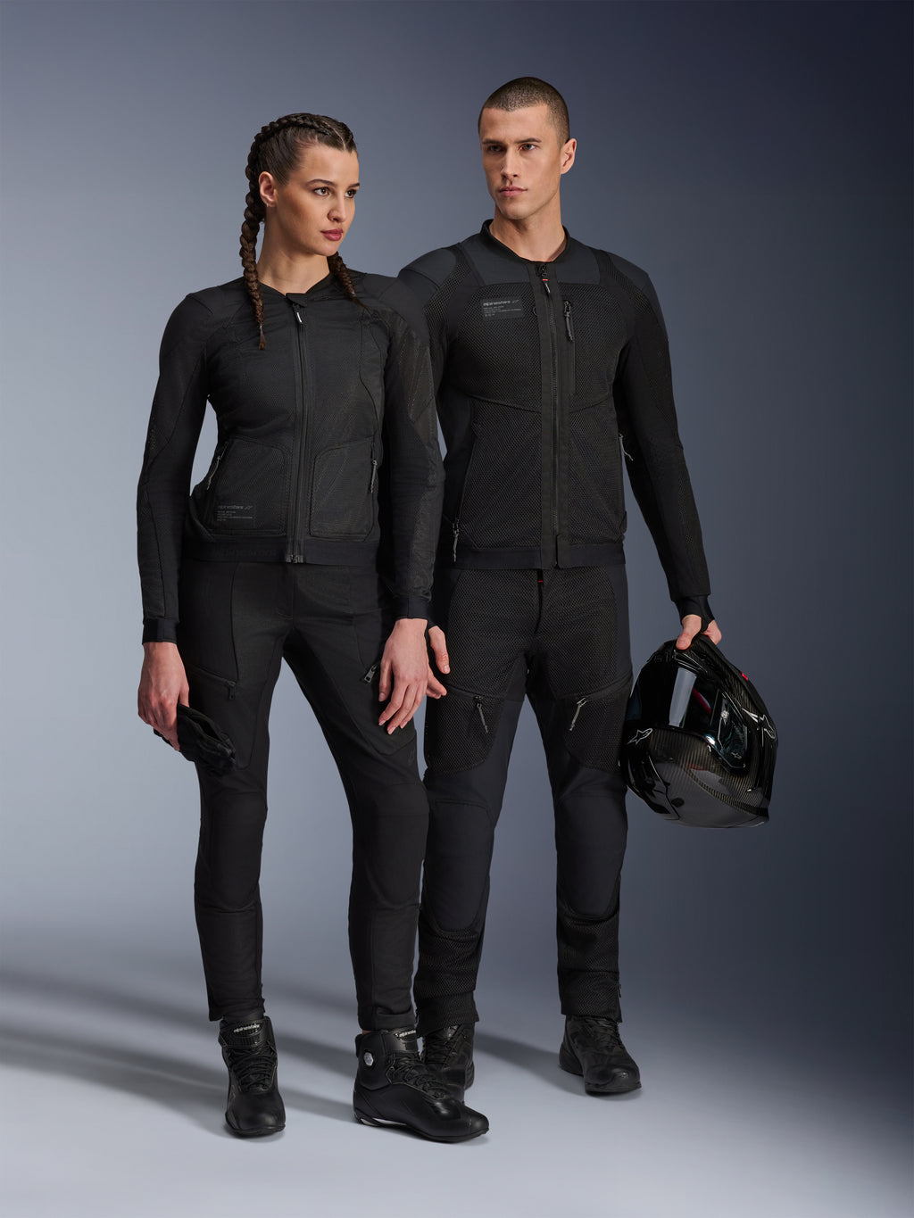 Alpinestars Stella Troop-Air Jacket, uomo e donna con abbigliamento tecnico da moto coordinato tutto nero, caratterizzato da struttura in mesh per la ventilazione, pantaloni e stivali neri, uomo con casco nero, abbigliamento per guida urbana.