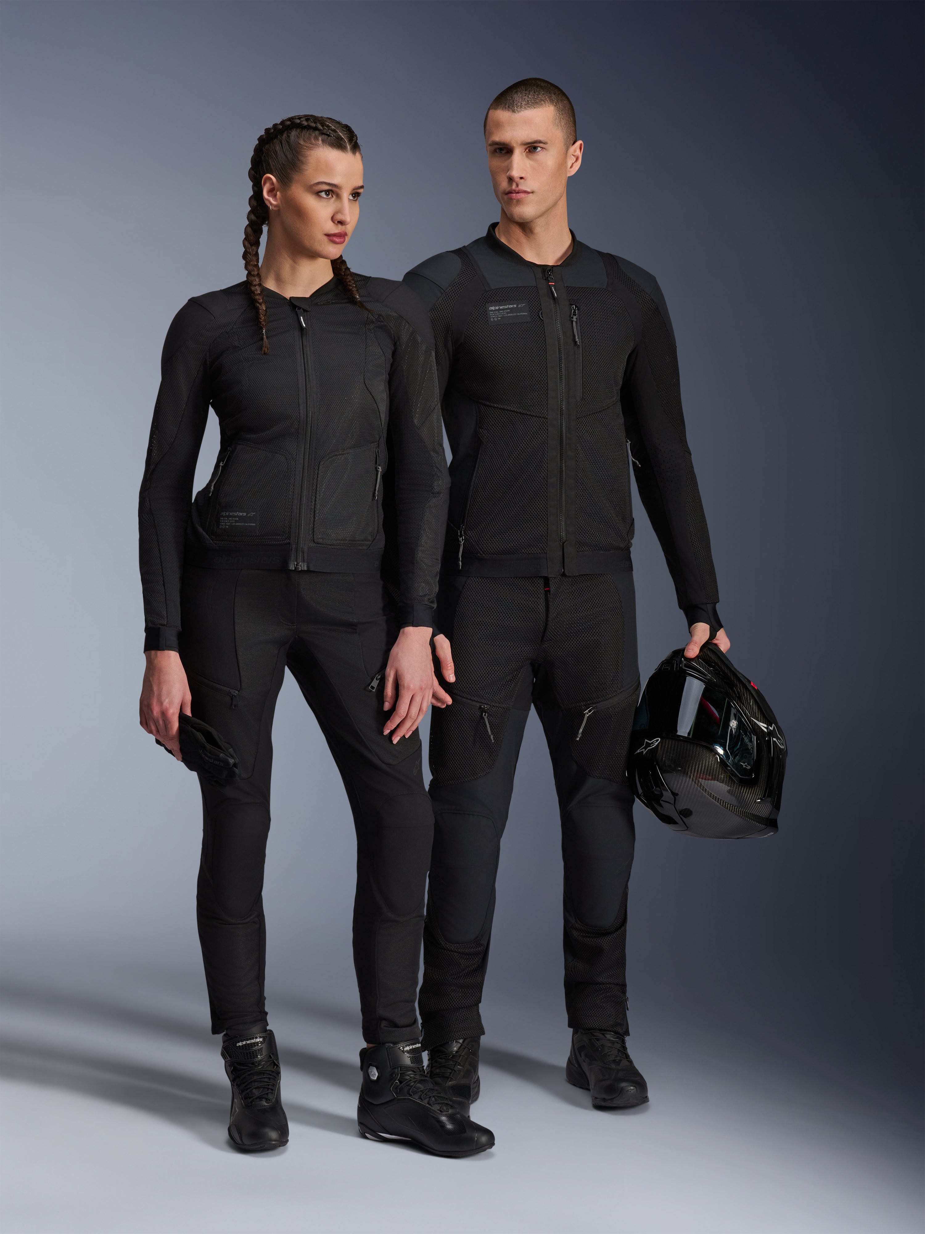 Alpinestars Stella Troop-Air Jacket, uomo e donna con abbigliamento tecnico da moto coordinato tutto nero, caratterizzato da struttura in mesh per la ventilazione, pantaloni e stivali neri, uomo con casco nero, abbigliamento per guida urbana.
