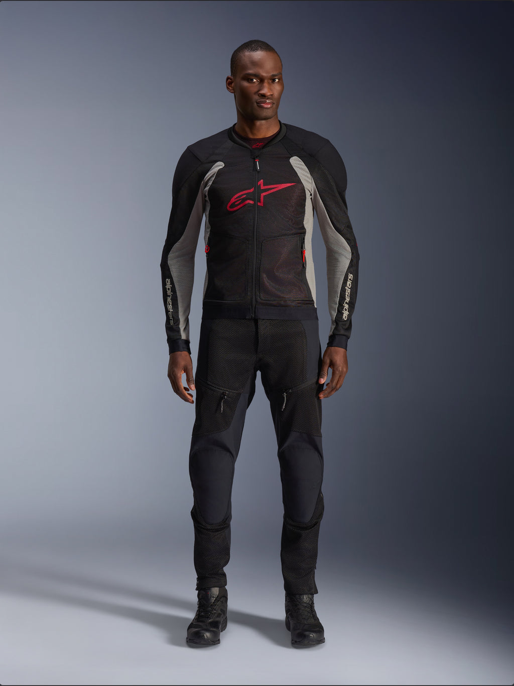 Modello che indossa la giacca Alpinestars Troop-Air Jacket in Black Ash Gray e Bright Red, giacca da moto tecnica in mesh con un evidente logo rosso sul petto, abbinata a pantaloni e stivali da moto neri, abbigliamento per la guida urbana.