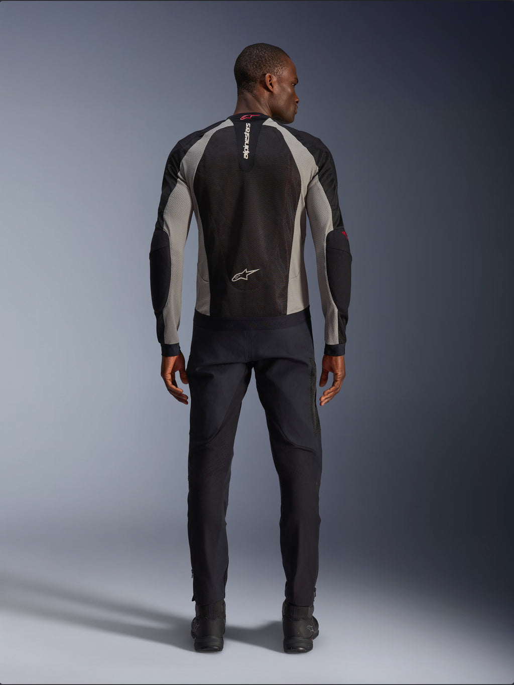 Giacca Alpinestars Troop-Air, giacca da moto nera, grigio cenere e rosso acceso, vista posteriore su modello, costruzione in mesh leggero per la ventilazione, logo Alpinestars sul colletto e sulla parte bassa della schiena, abbinata a pantaloni da moto neri