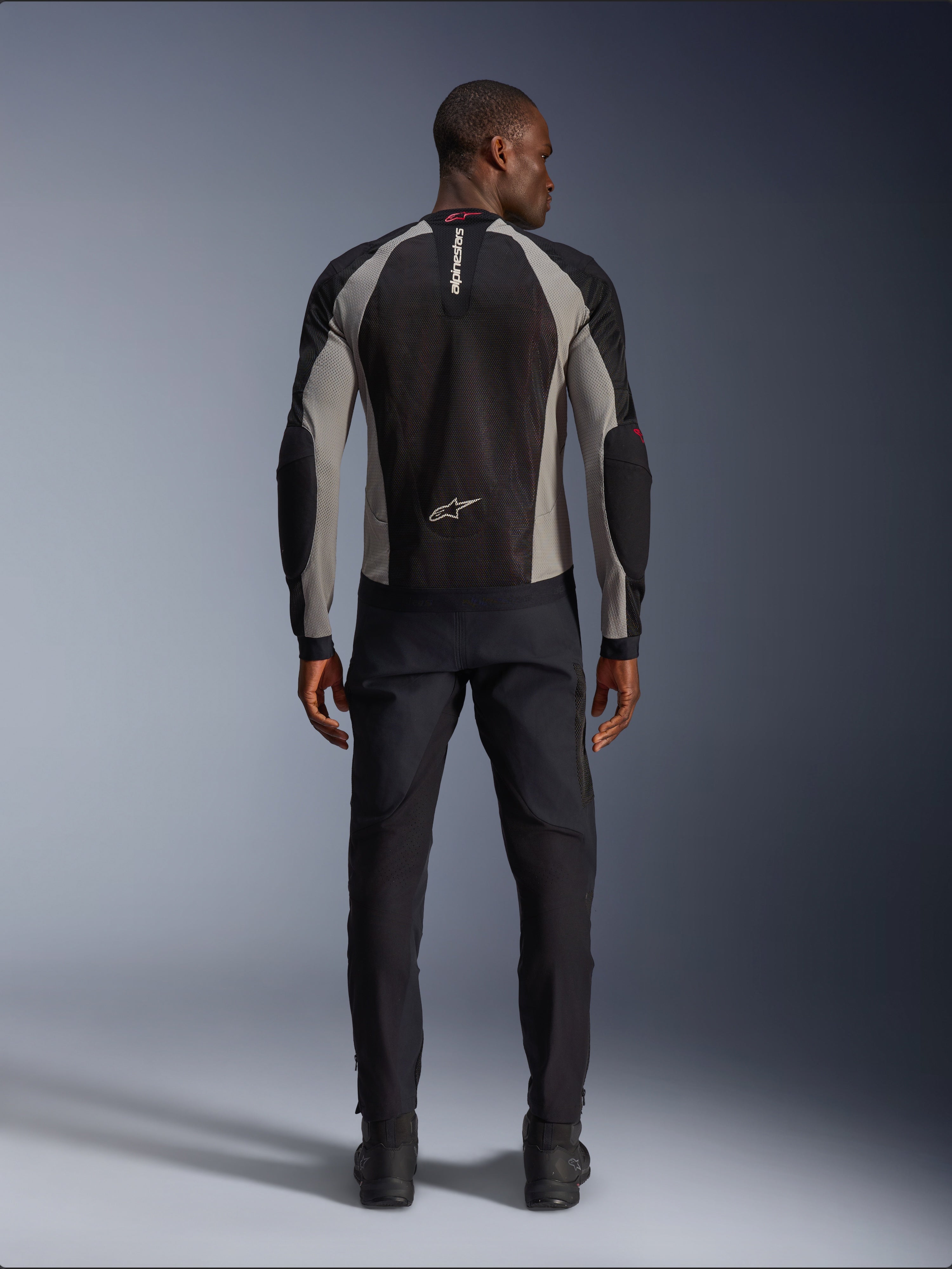 Giacca Alpinestars Troop-Air, giacca da moto nera, grigio cenere e rosso acceso, vista posteriore su modello, costruzione in mesh leggero per la ventilazione, logo Alpinestars sul colletto e sulla parte bassa della schiena, abbinata a pantaloni da moto neri