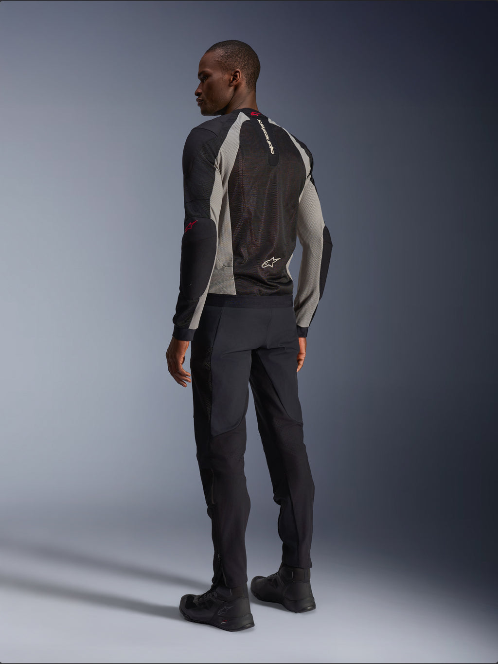 Modello che indossa la giacca Alpinestars Troop-Air vista da dietro, nero e grigio cenere con accenti rosso brillante, costruzione in mesh tecnica per la ventilazione, abbinata a pantaloni da moto neri e stivali da guida, stile urban contemporaneo