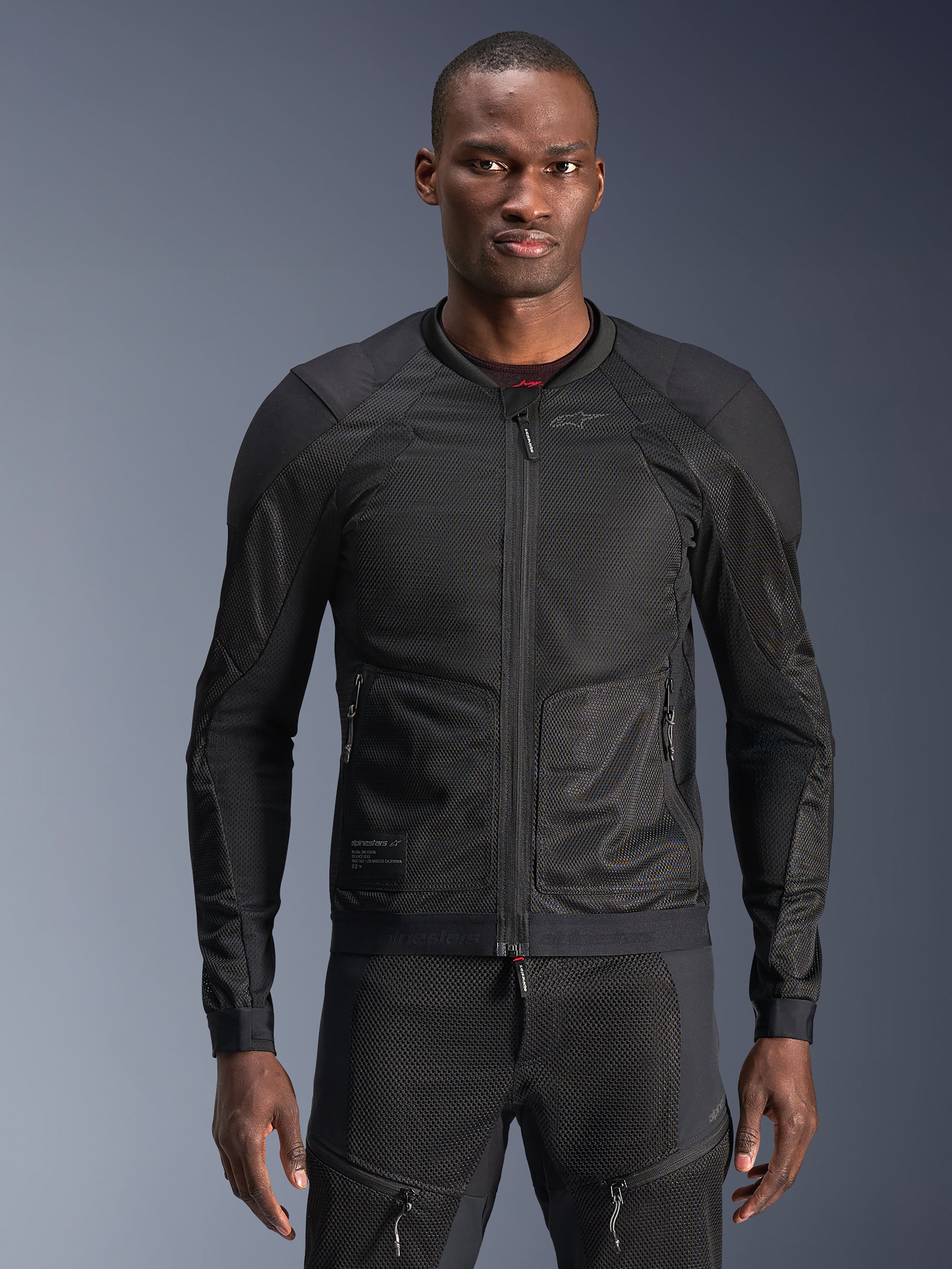 Giacca Alpinestars Troop-Air, giacca da moto in mesh nera, indossata da un modello con pantaloni in mesh neri coordinati, struttura in mesh tecnico 2D traspirante con pannelli rinforzati sulle spalle, cerniera frontale, abbigliamento da moto urbano moderno