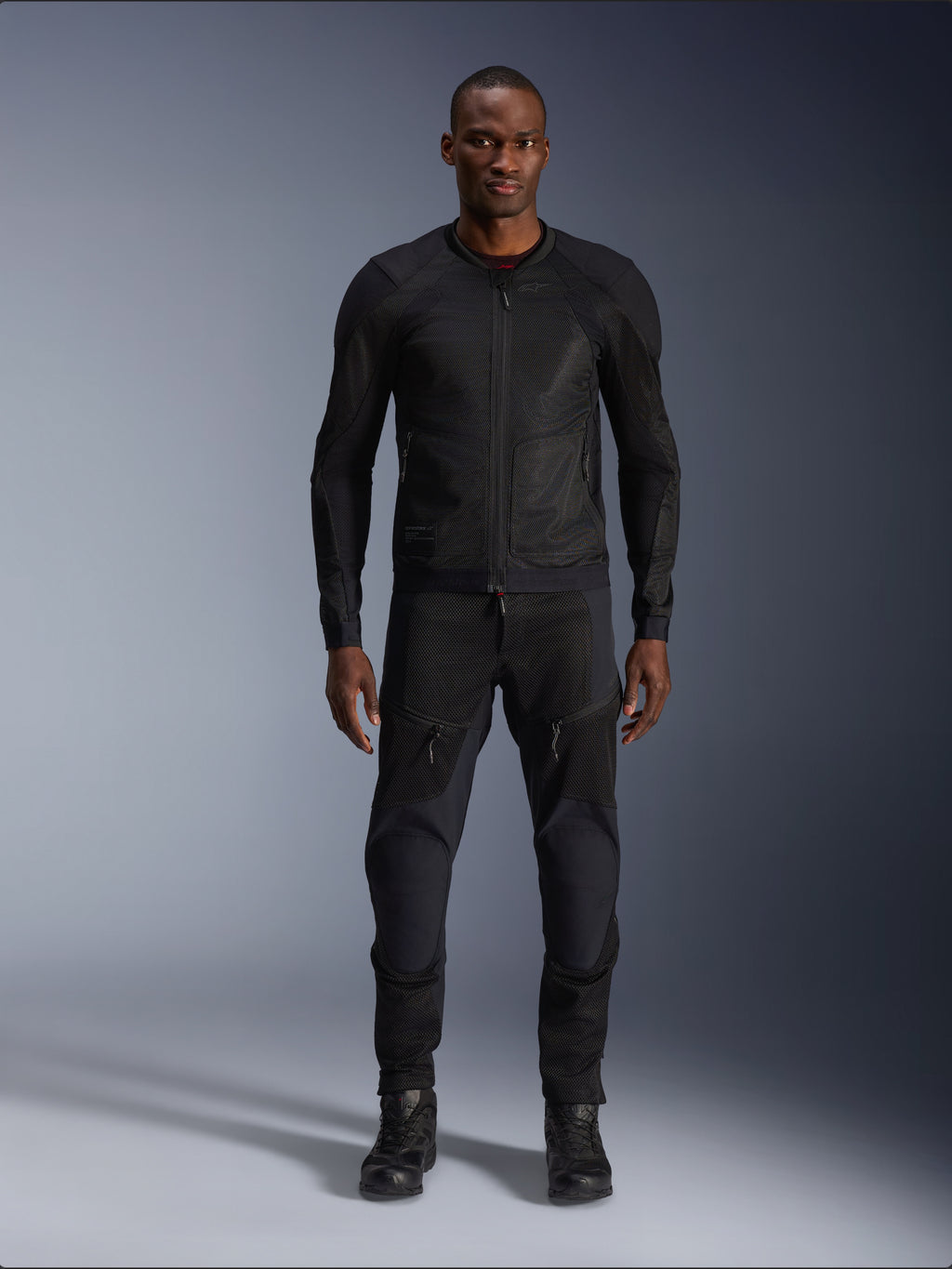 Modello uomo che indossa la giacca Alpinestars Troop-Air in Black Black, giacca da moto in mesh leggero, abbinata a pantaloni da moto e stivali neri coordinati, caratterizzata da una struttura in mesh tecnologico 2D per un flusso d'aria superiore.