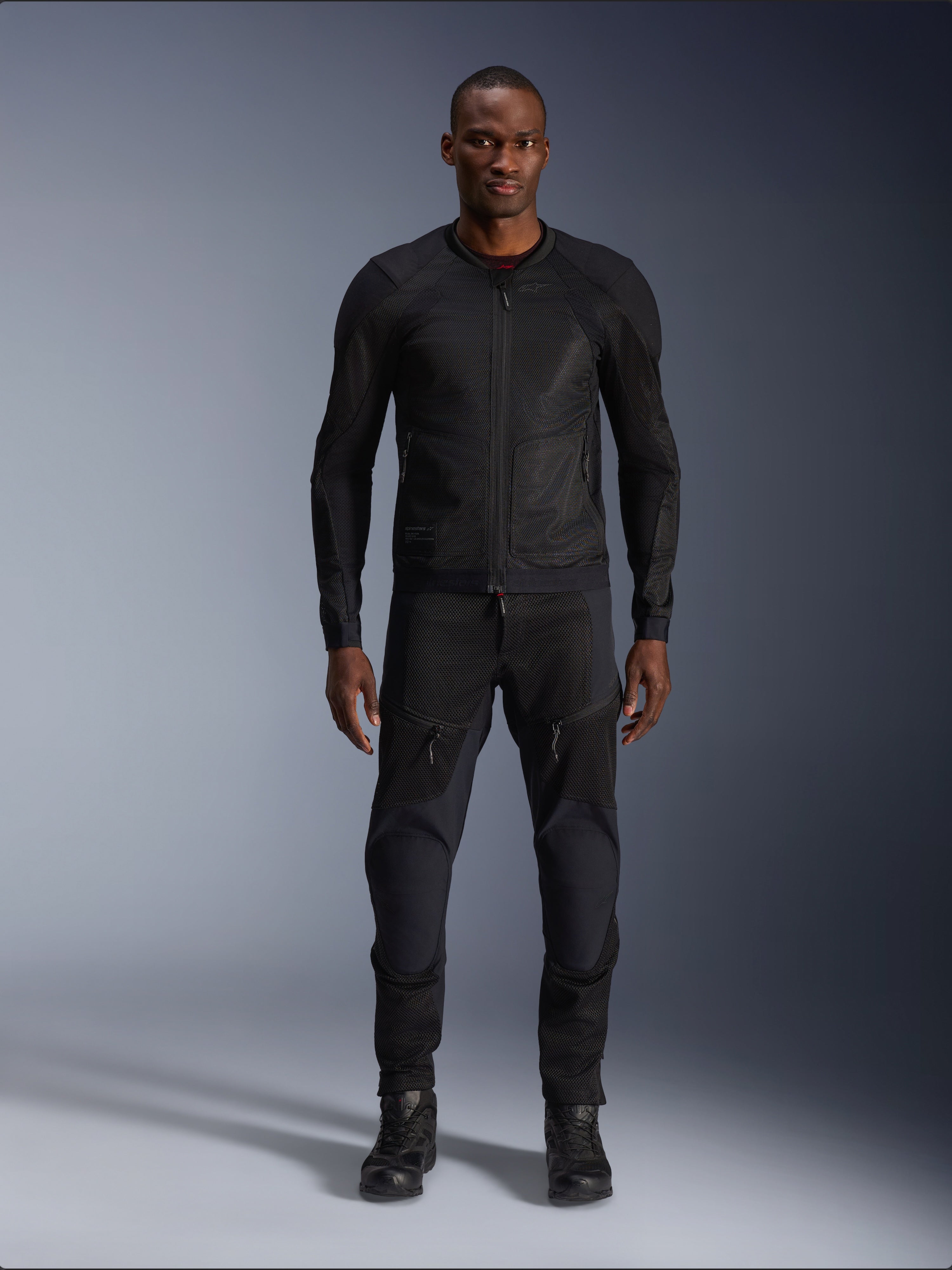 Modello uomo che indossa la giacca Alpinestars Troop-Air in Black Black, giacca da moto in mesh leggero, abbinata a pantaloni da moto e stivali neri coordinati, caratterizzata da una struttura in mesh tecnologico 2D per un flusso d'aria superiore.