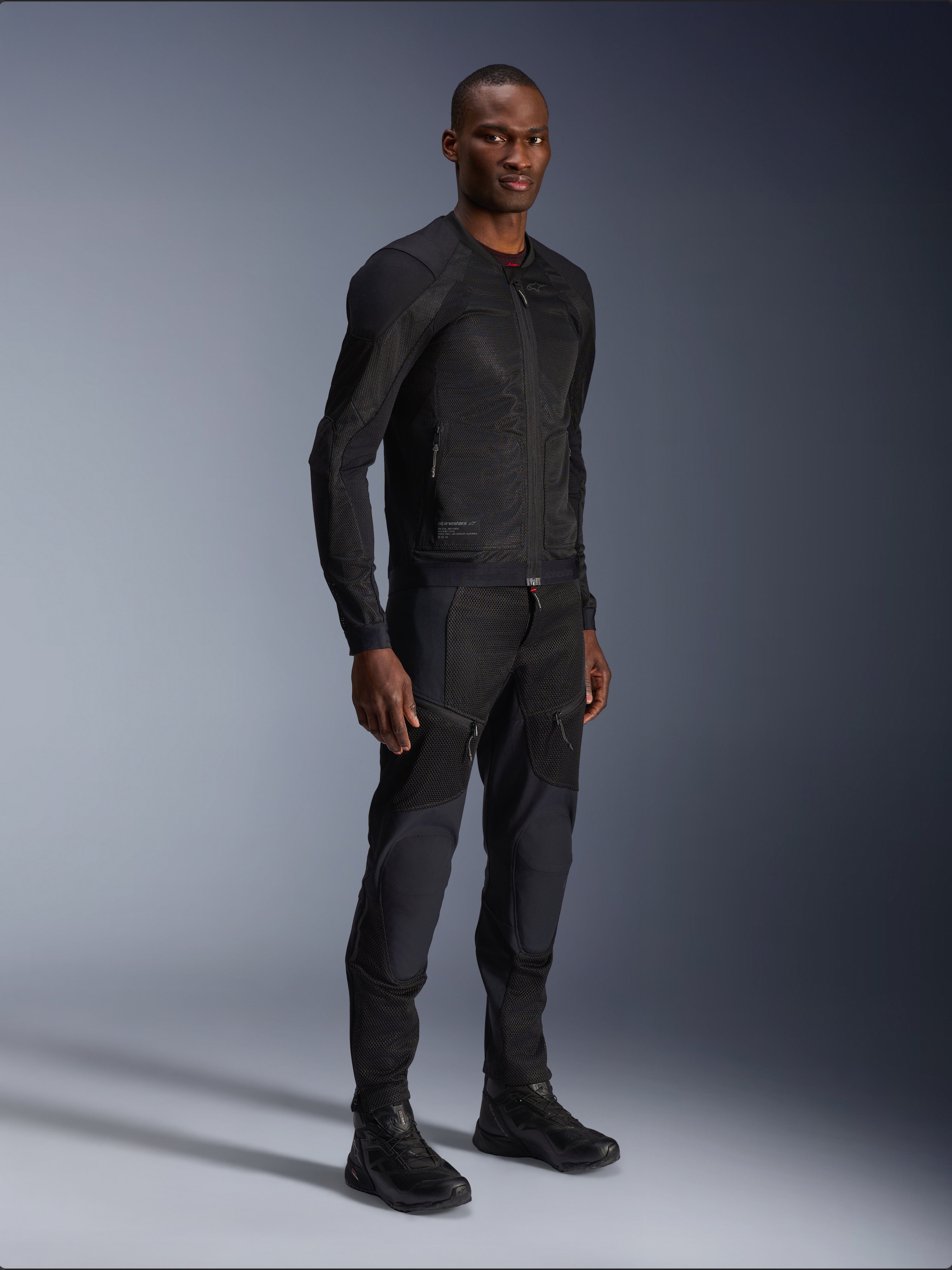 Alpinestars Troop-Air Jacket, nera, giacca da moto in mesh tecnico indossata da un modello con pantaloni e stivali neri coordinati, con struttura in mesh leggero e design urbano minimalista