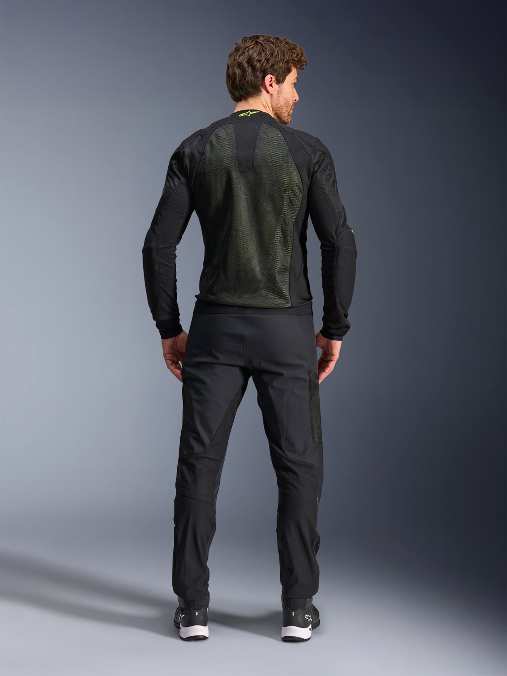 Giacca Alpinestars Troop-Air, Nero Giallo Fluo, vista posteriore su un modello, con ampio pannello posteriore in mesh tecnico 2D per la ventilazione, giacca tecnica da moto indossata con pantaloni da moto e stivali neri, stile motociclistico urbano