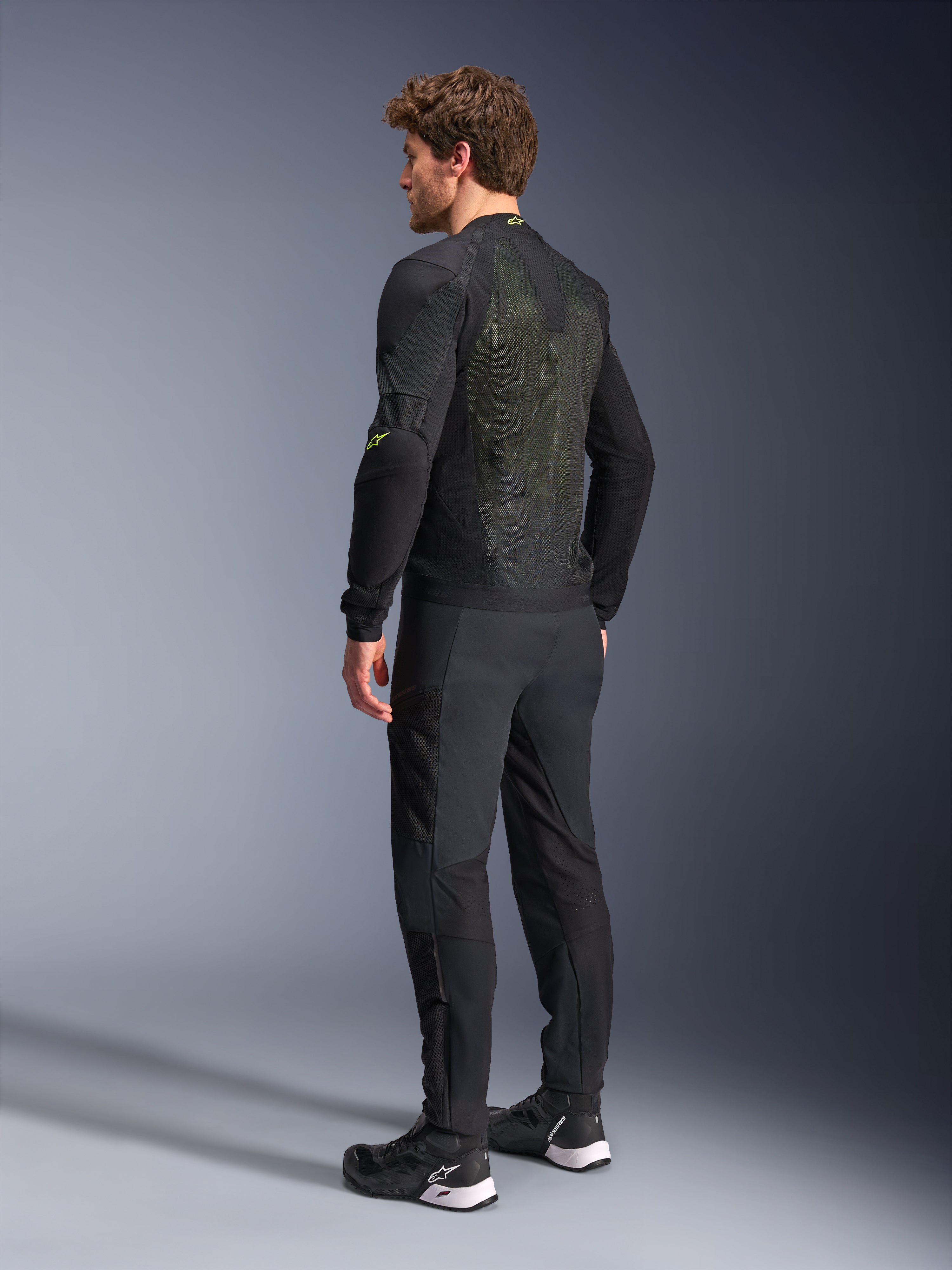 Giacca Alpinestars Troop-Air in nero e giallo fluo, vista posteriore di un modello che indossa la giacca tecnica, ampio pannello posteriore in mesh con riflessi gialli e loghi Alpinestars giallo fluo, abbinata a pantaloni da moto neri