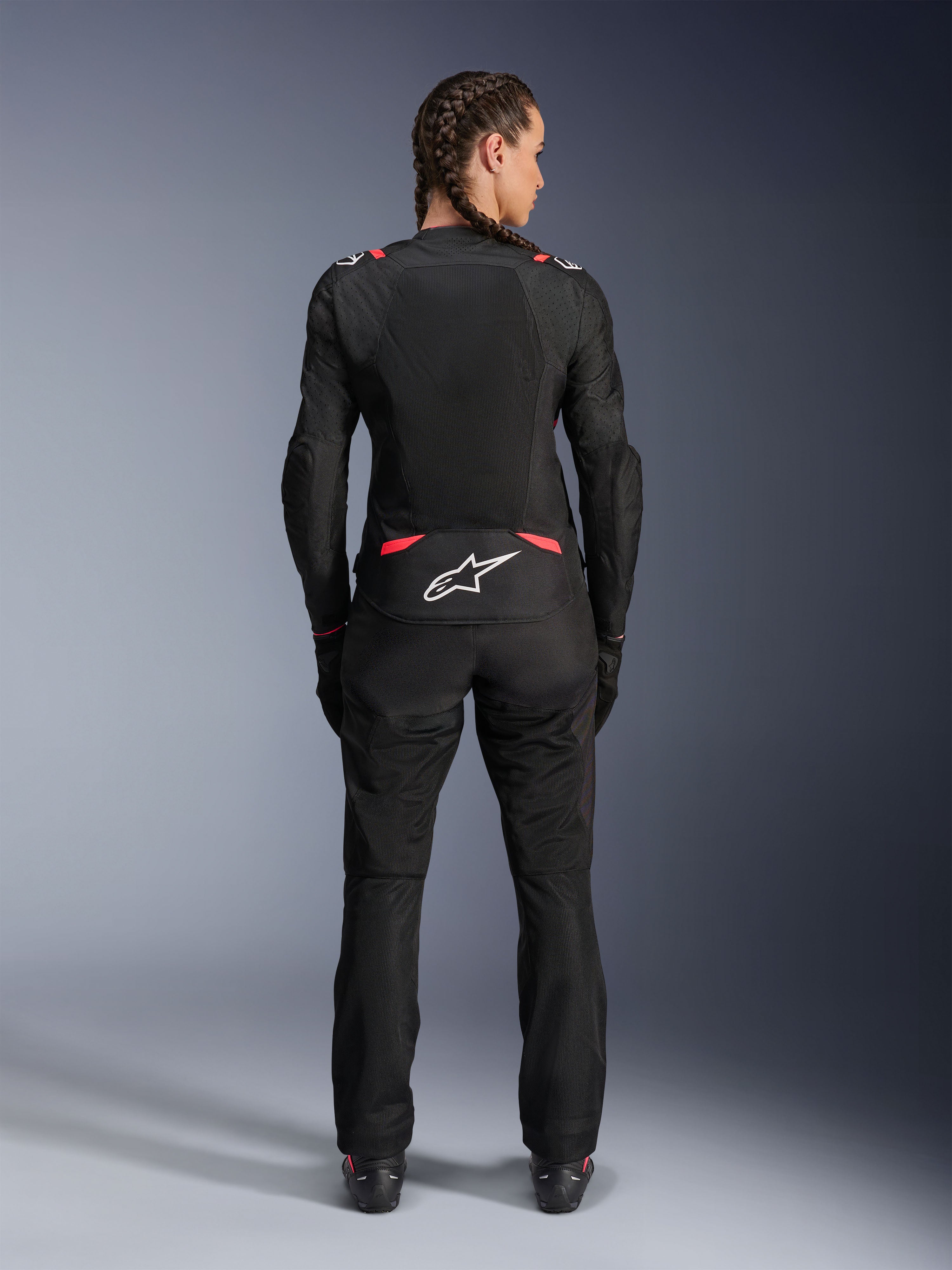 Alpinestars Stella T-SPS Air V2 Jacket, giacca da moto in tessuto da donna, nero e rosa Diva, vista posteriore su modella con capelli intrecciati, con ampi pannelli in mesh per la ventilazione, logo Astars bianco sulla parte inferiore della schiena, abbinata a pantaloni da moto neri coordinati.