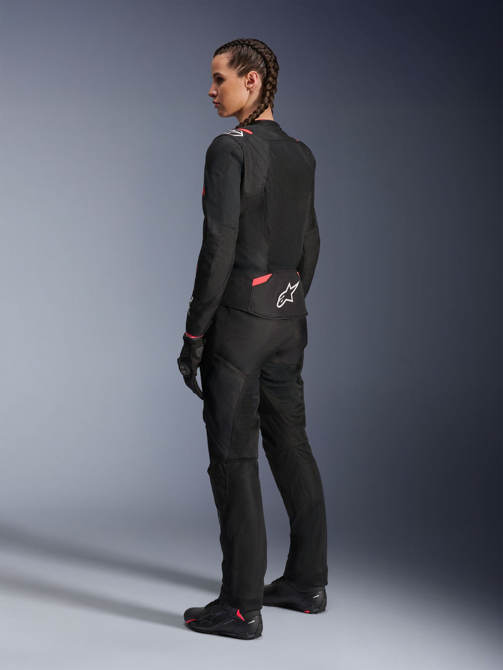 Giacca Alpinestars Stella T-Sps Air v2 da donna, nero e Diva Pink, giacca da moto in tessuto vista da una prospettiva posteriore-laterale, con ampi pannelli in mesh per il flusso d'aria e logo a stella bianco sulla schiena, indossata con pantaloni da moto, guanti e stivali coordinati neri.