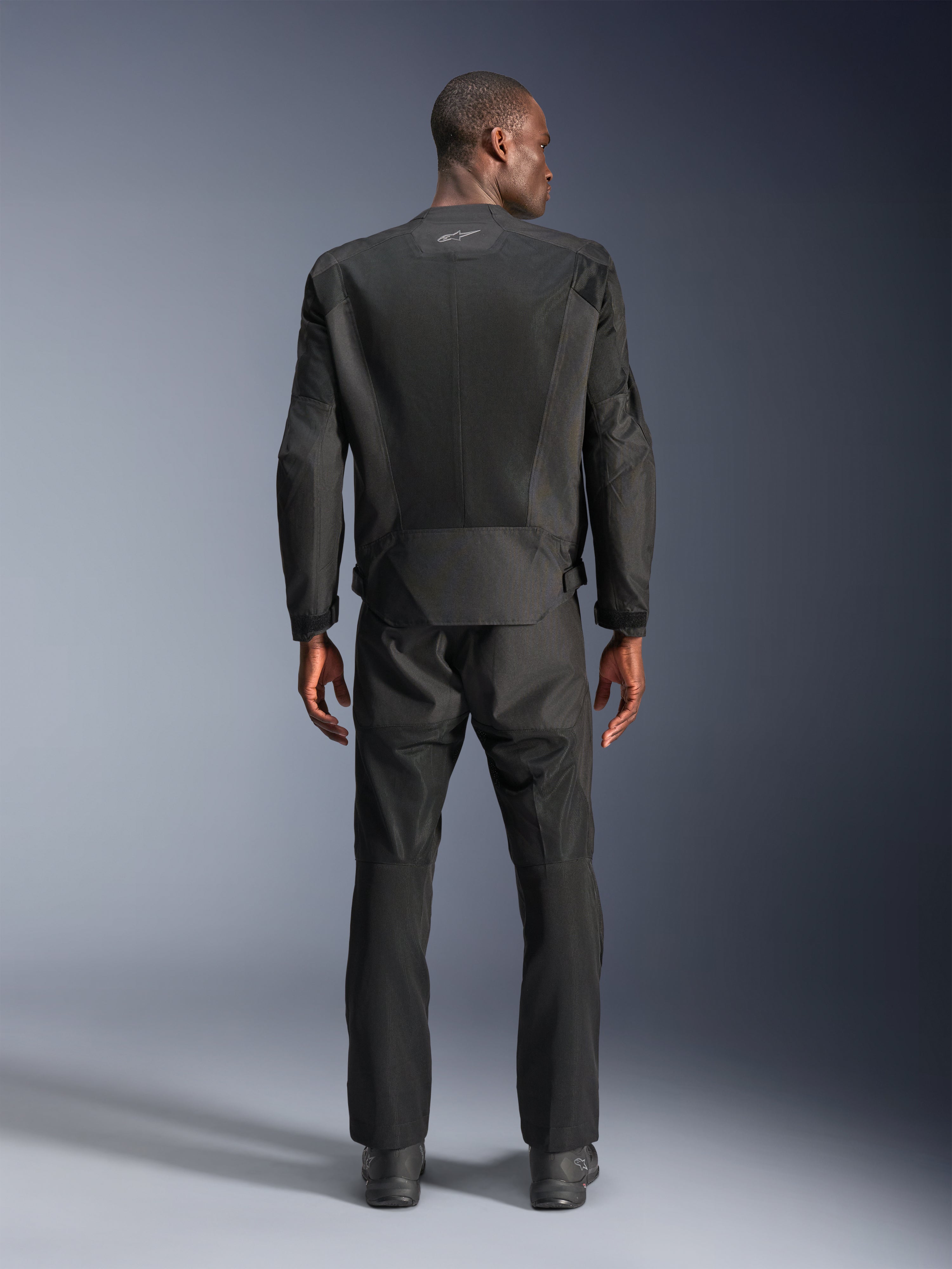 Vista posteriore di un uomo che indossa la giacca Alpinestars C-1 Air in nero, giacca da moto urbana con pannelli in rete tecnica, pantaloni in tessuto e stivali neri coordinati, estetica pulita con logo riflettente sulla parte superiore della schiena.