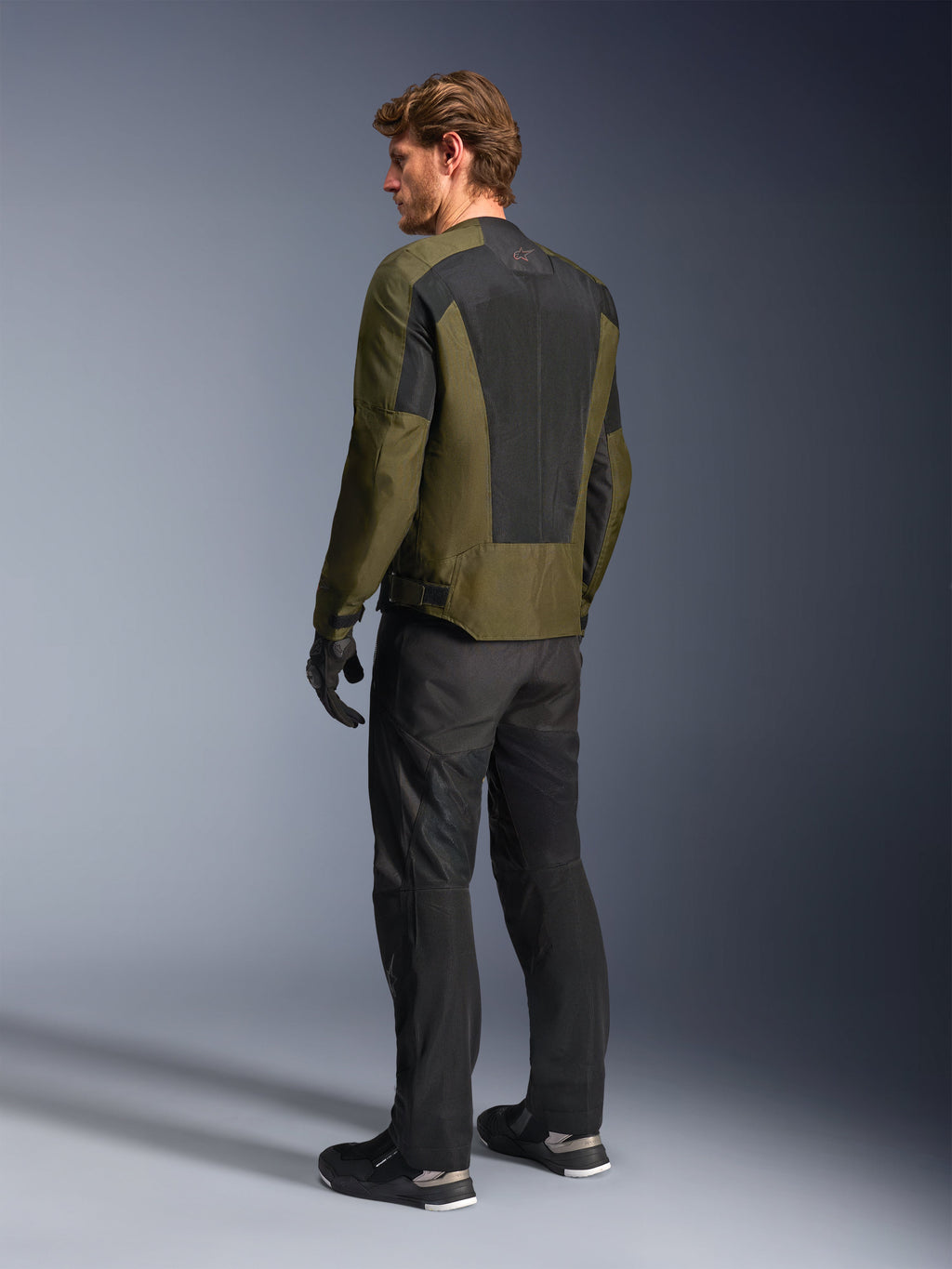 Modello uomo con giacca Alpinestars C-1 Air in colore Khaki, giacca da moto urban, vista posteriore con ampi pannelli in rete nera sulla schiena, abbinata a pantaloni tecnici da guida e guanti neri, abbigliamento da moto contemporaneo per climi caldi