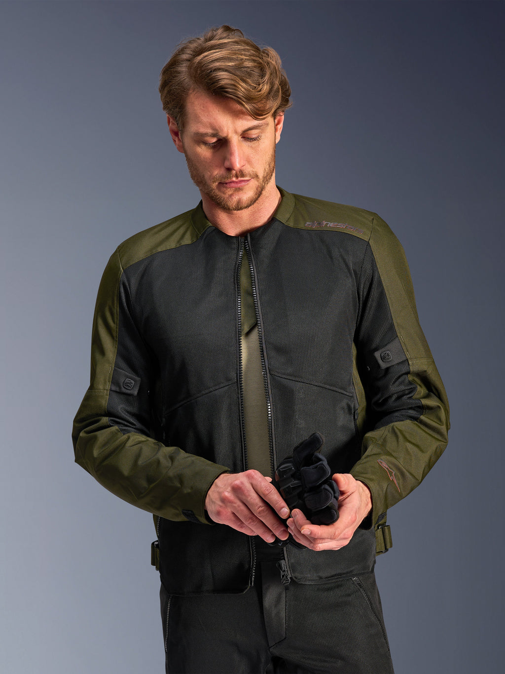 Uomo che indossa la giacca Alpinestars C-1 Air in Khaki, giacca da moto urban con tessuto verde oliva e pannelli in rete nera, mentre guarda in basso tenendo dei guanti da moto neri, stile pulito e contemporaneo per la guida su strada.