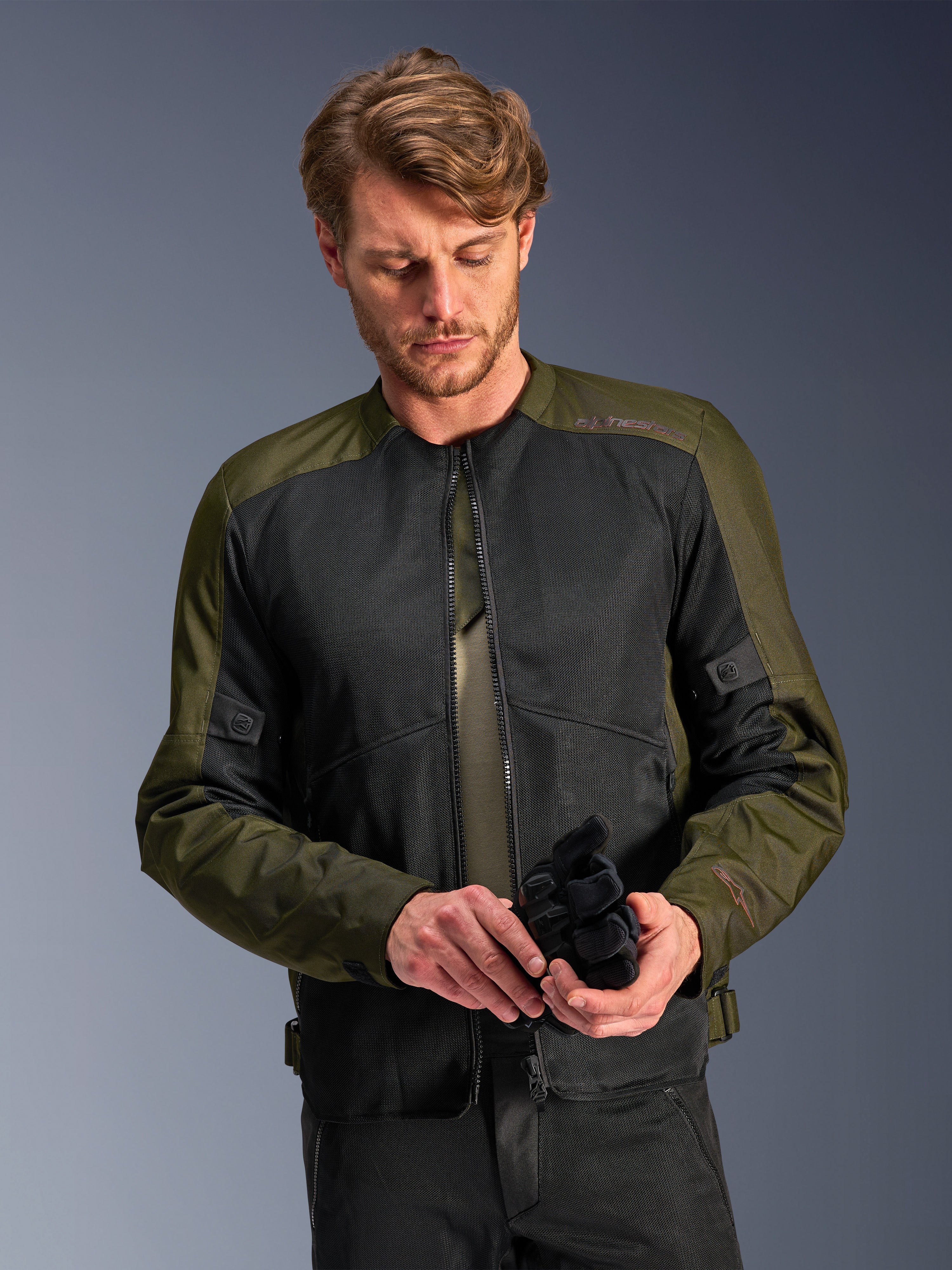 Uomo che indossa la giacca Alpinestars C-1 Air in Khaki, giacca da moto urban con tessuto verde oliva e pannelli in rete nera, mentre guarda in basso tenendo dei guanti da moto neri, stile pulito e contemporaneo per la guida su strada.