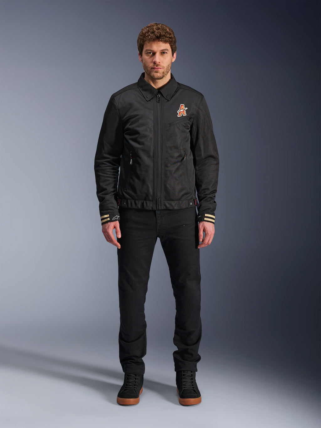 Alpinestars Ignite Air Jacket, giacca da moto nera con colletto a camicia, cerniera frontale e logo arancione sul petto, indossata da un modello con pantaloni e sneakers nere per un look urban contemporary.