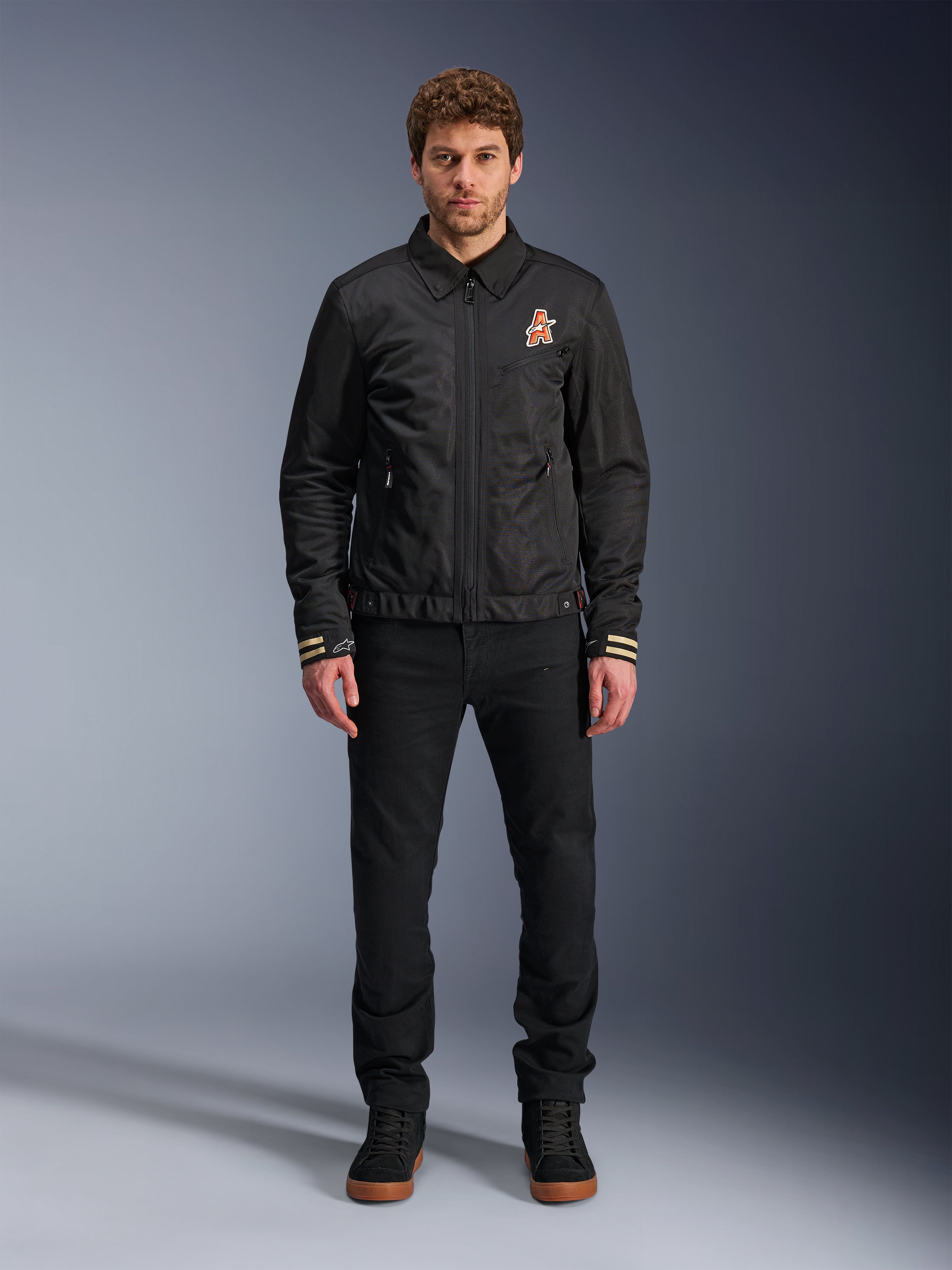 Alpinestars Ignite Air Jacket, giacca da moto nera con colletto a camicia, cerniera frontale e logo arancione sul petto, indossata da un modello con pantaloni e sneakers nere per un look urban contemporary.