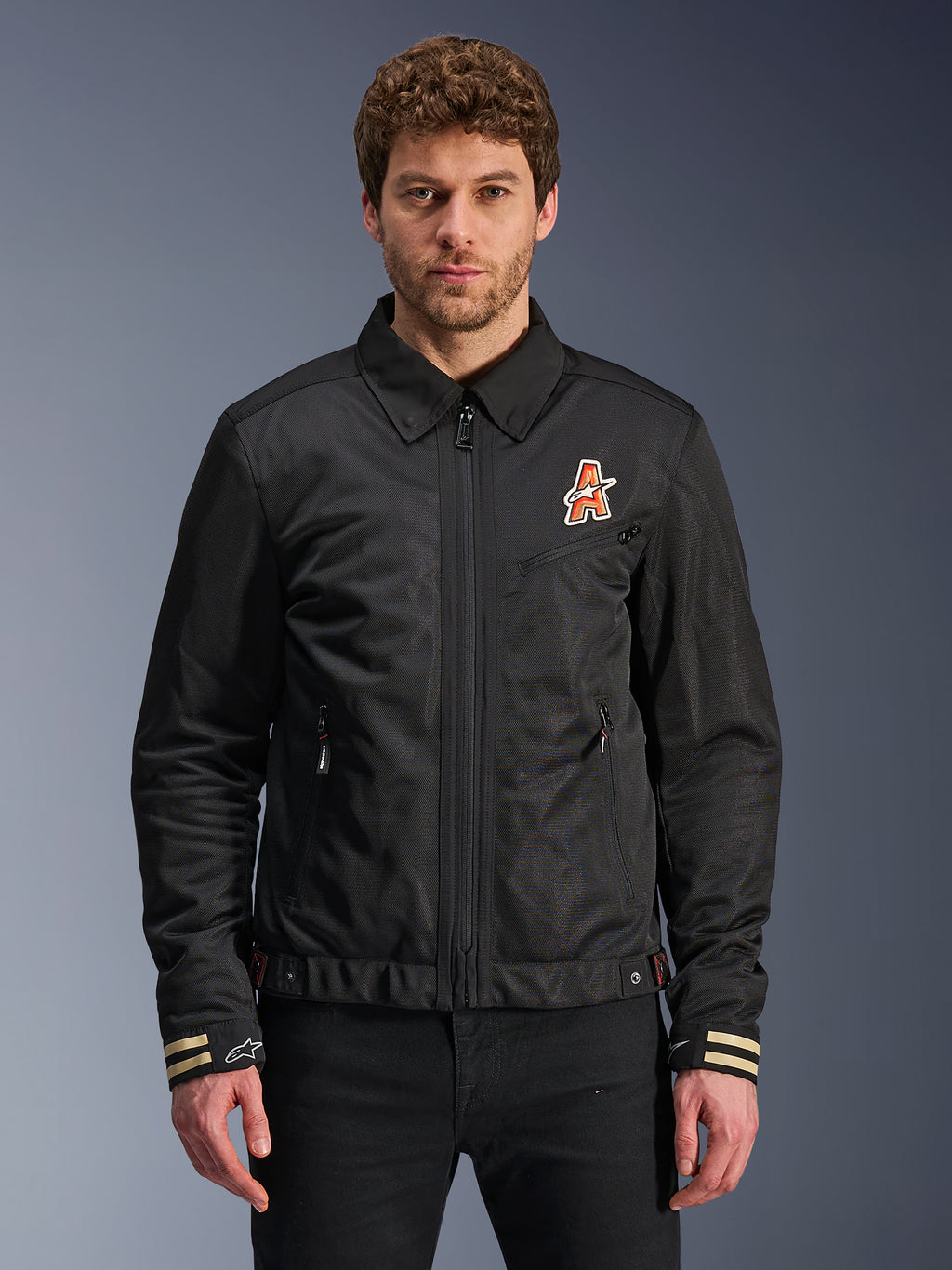 Alpinestars Ignite Air Jacket, giacca da moto nera, colletto tradizionale a camicia, logo a 'A' arancione e bianco sul petto, polsini a strisce beige con logo a stella bianco, abbigliamento da moto urban contemporary