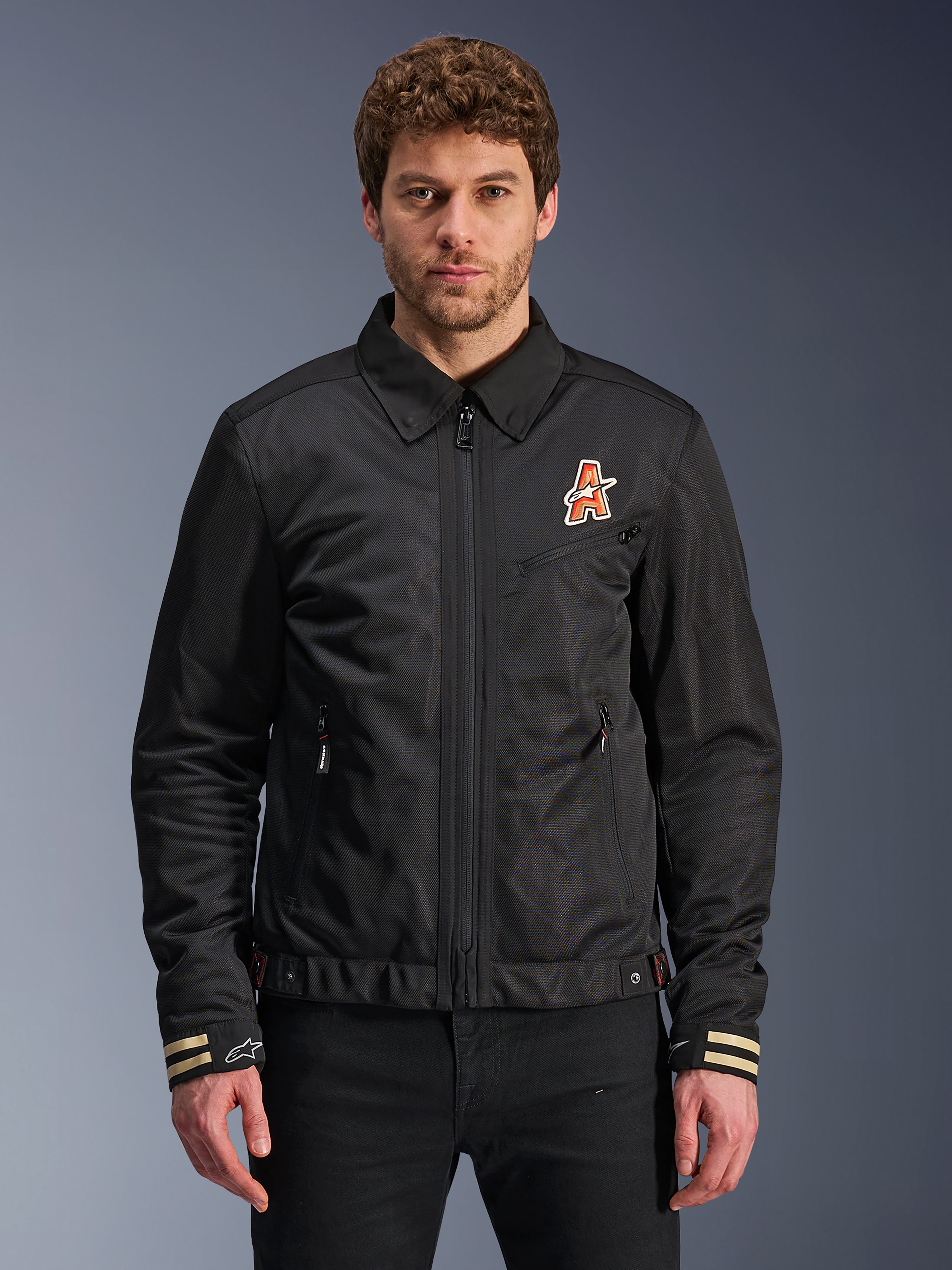 Alpinestars Ignite Air Jacket, giacca da moto nera, colletto tradizionale a camicia, logo a 'A' arancione e bianco sul petto, polsini a strisce beige con logo a stella bianco, abbigliamento da moto urban contemporary