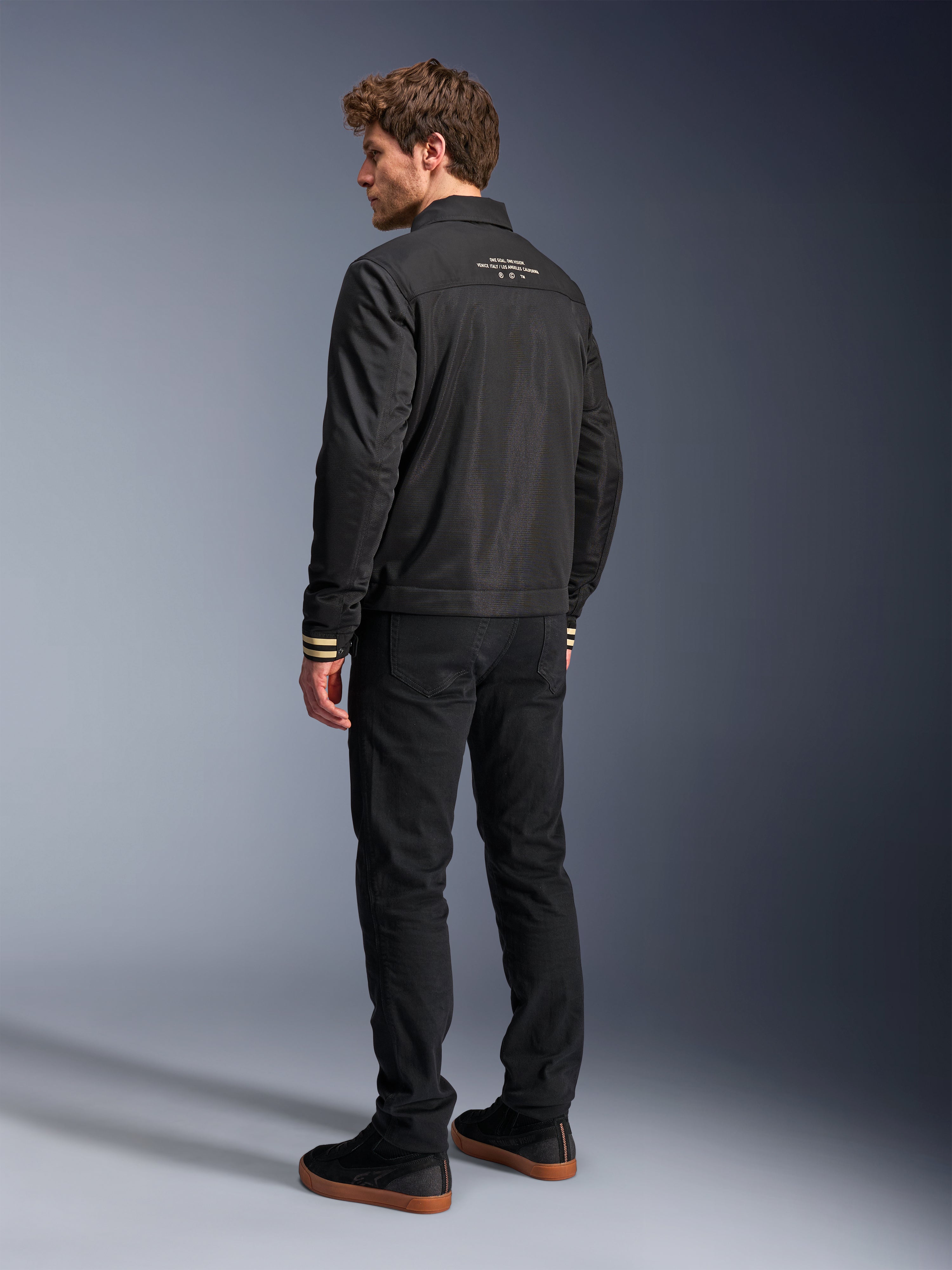 Giacca Alpinestars Ignite Air, giacca da moto nera, vista posteriore con testo grafico bianco sulla parte superiore della schiena e polsini a righe beige, silhouette stile Harrington con corpo in rete, abbinata a pantaloni neri e sneakers nere